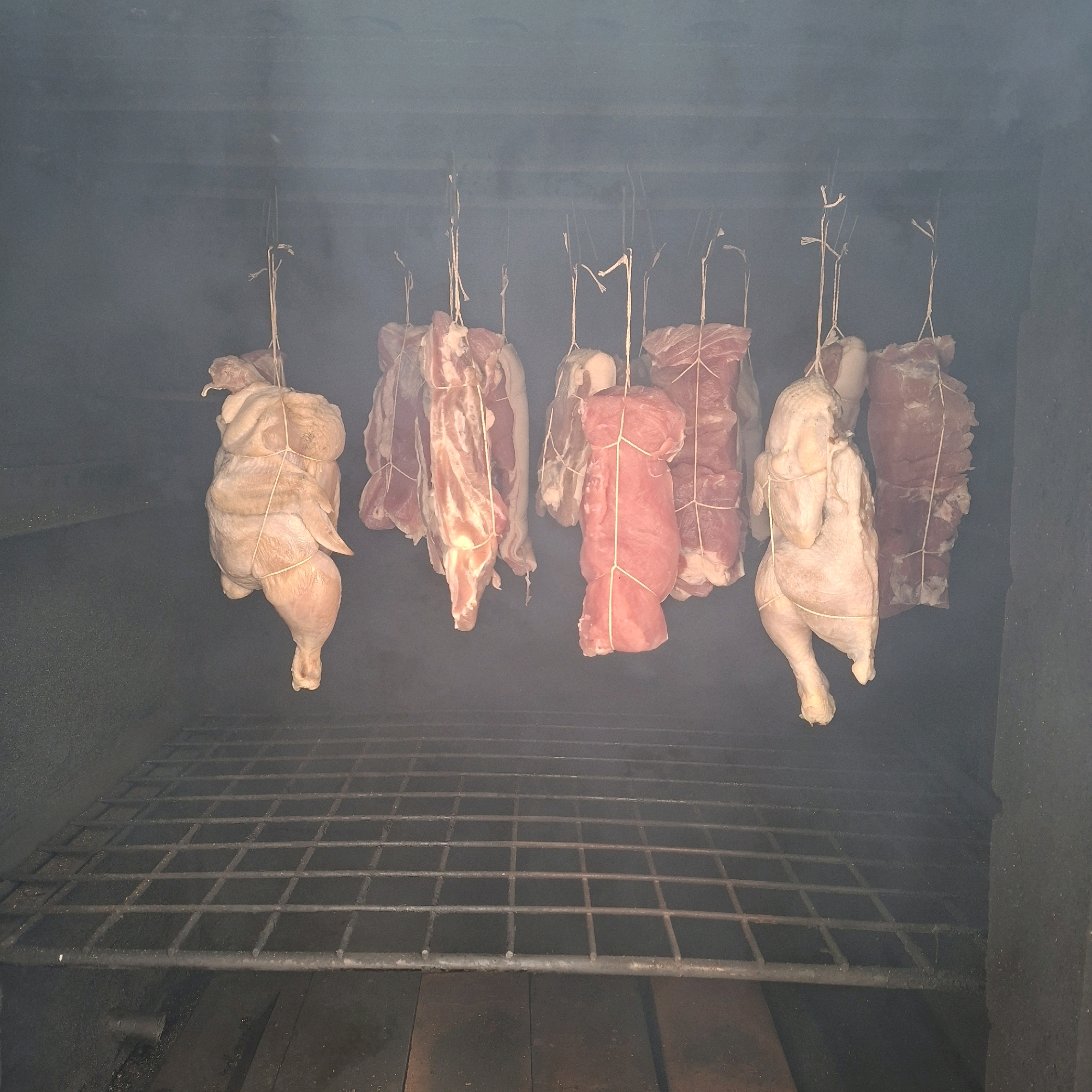 A raw chicken and several cuts of raw meat are suspended by twine inside a dark, smoky enclosure, likely a smoker. The chicken is whole and trussed. The meat cuts vary in shape and size, with some appearing to be pork roasts and others possibly bacon. They are hung in a line, with a metal grate positioned below them, suggesting they are being prepared for smoking. The environment appears industrial or rustic, with dark walls and a wooden floor beneath the grate. There is a noticeable haze of smoke throughout the enclosure. The location of Anapskaya, Russia is relevant to the context of the image.