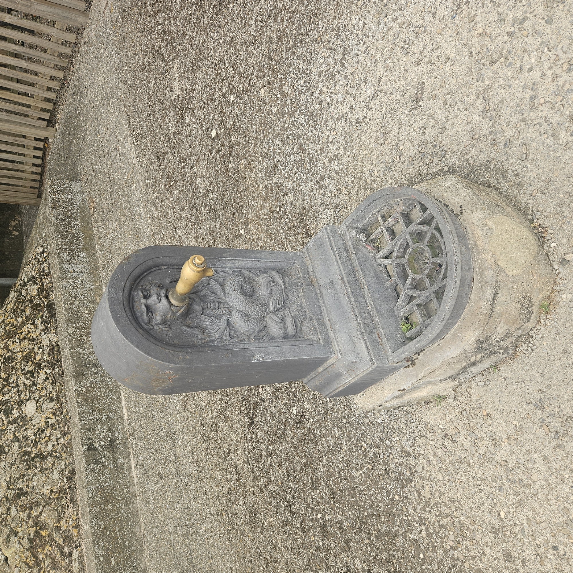 A weathered, dark gray cast iron public fountain is centered in the frame, mounted on a rough concrete base. The fountain's facade features an engraved relief of a grotesque human-like face with wide eyes and an open mouth. Extending from the mouth is a brass spout. Below the face, there is a relief of a scaled, serpentine creature. The bottom of the fountain has a circular, ornate drain grate, surrounded by a shallow basin. Small green weeds are visible growing within the grate and around the base. The fountain is situated on a rough, gravelly surface. In the background, a rough concrete wall with stone debris is visible on the left, and a weathered wooden fence runs along the right side of the frame. The overall setting appears to be an outdoor public area, possibly in Aiguaviva, Spain, though this cannot be definitively confirmed from the image alone. There are no people or animals present.