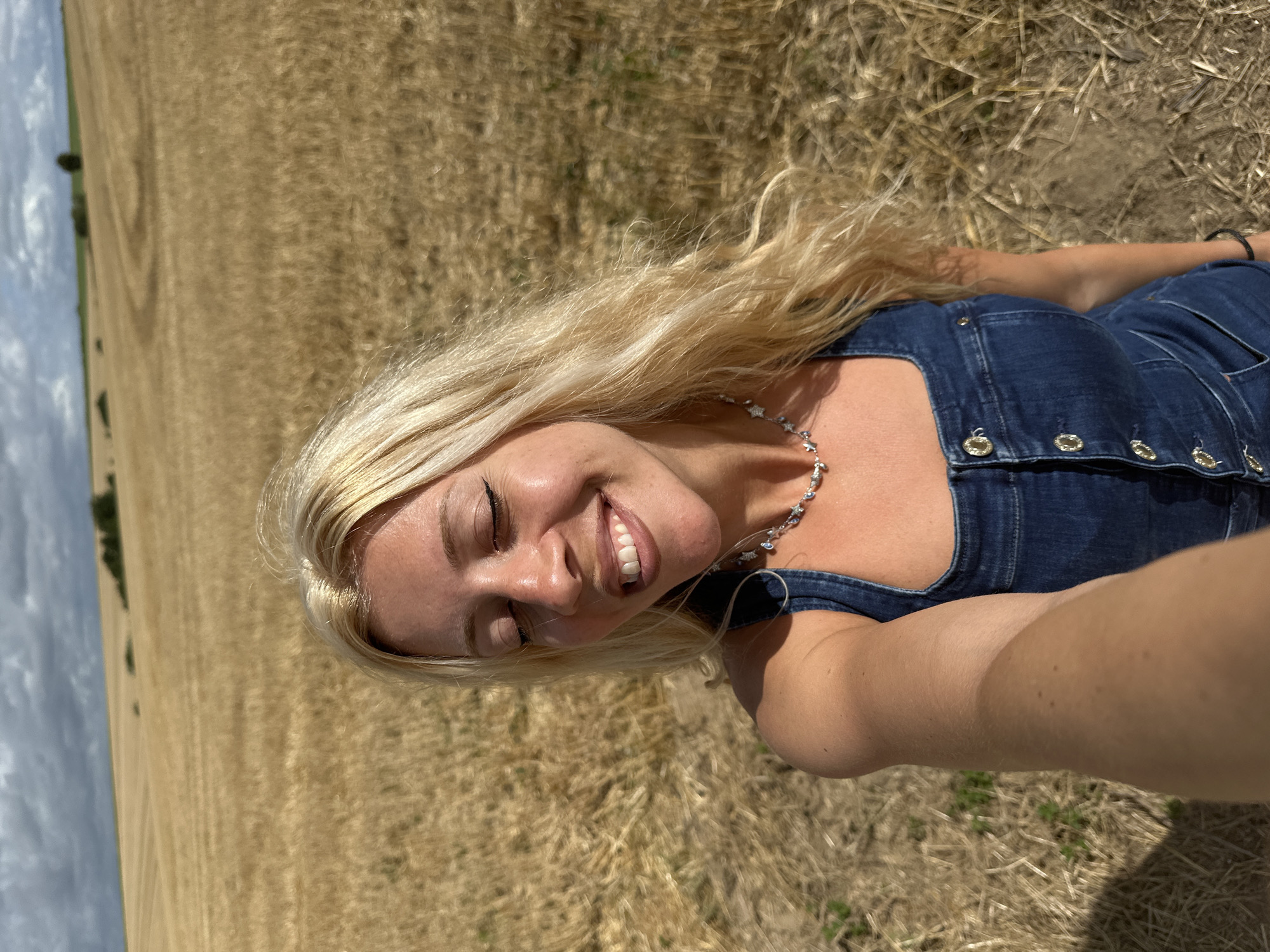 A young woman with blonde hair and closed eyes smiles in a selfie taken in a dry, golden field under a cloudy sky. She is wearing a denim jumpsuit and a beaded necklace. The field appears to be a harvested crop field, possibly wheat or barley, with stubble covering the ground. In the distance, there are rolling hills with some scattered trees. The overall setting suggests an outdoor, rural environment. The location is identified as Royon, France.