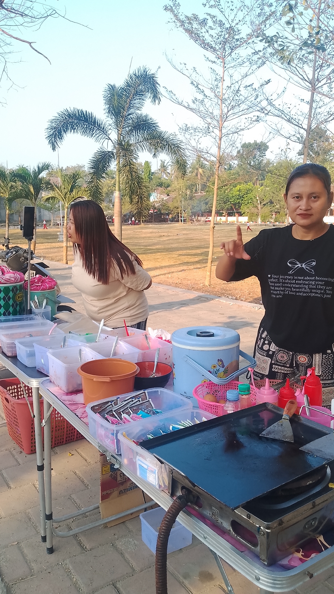 In Karanglangu, Indonesia, two women are operating a food stall in an outdoor setting. The stall is set up on a folding table laden with various food ingredients in plastic containers, bottles of condiments, and a griddle. A large blue water cooler is also present on the table. One woman, on the right, wears a black t-shirt with a white quote and a patterned apron. She is facing the camera, gesturing with her right hand. The other woman, on the left, has long brown hair and is wearing a light-colored t-shirt. She is turned away from the camera, looking towards the left. The background shows a grassy field with trees, including palm trees, and a clear sky, suggesting it is daytime. In the distance, there are some structures and activity, possibly indicating a public area. The ground is paved with interlocking bricks.