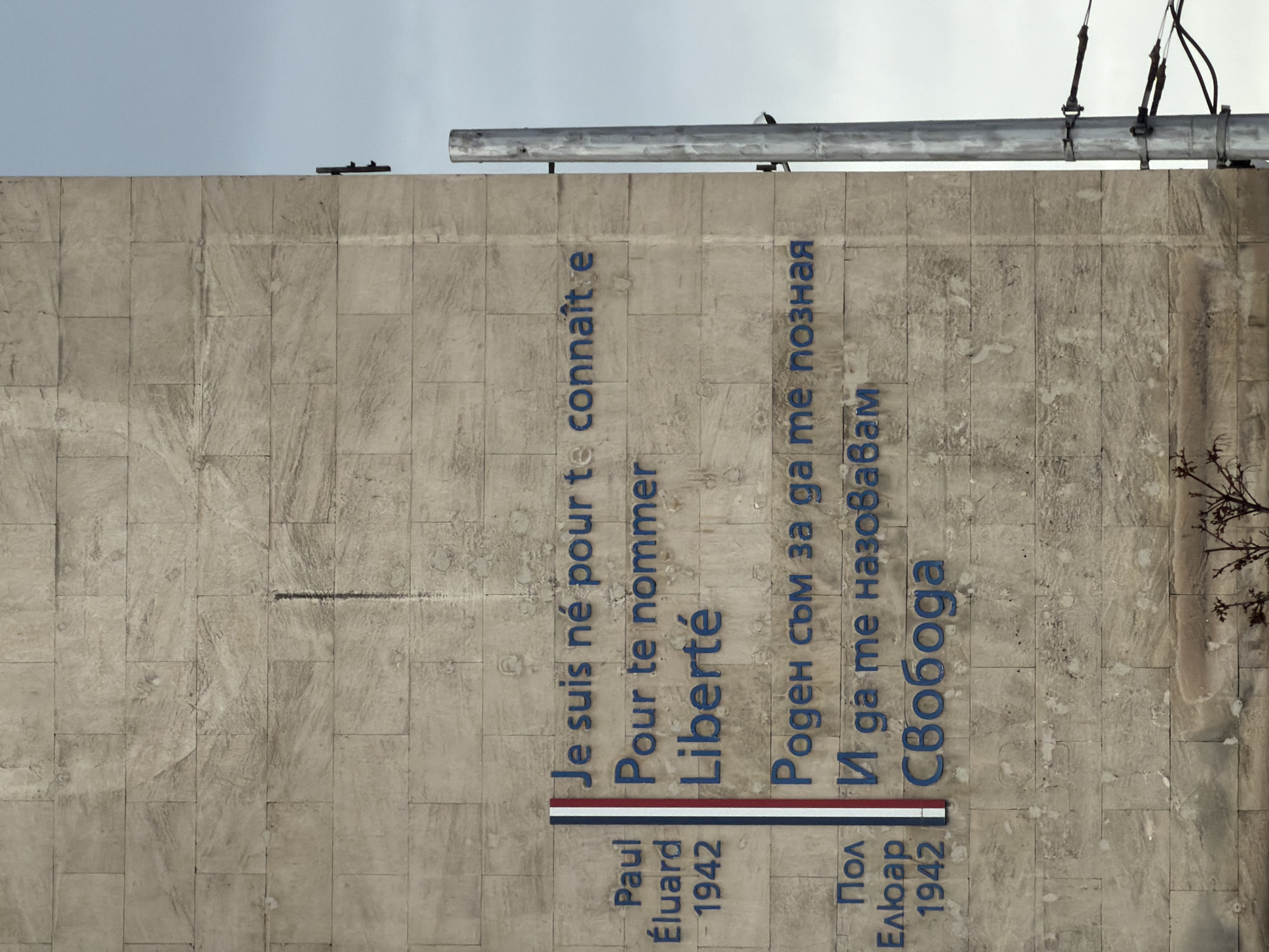 The image captures a stone monument in Sofia, Bulgaria, displaying text in both French and Bulgarian. On the left side of the monument, the French inscription reads, "Je suis né pour te connaître. Pour te nommer. Liberté." Underneath, it states "Paul Éluard 1942". A horizontal bar in the colors of the French flag is positioned between the text and the name. On the right side, the corresponding Bulgarian text says, "Роден съм за да те позная. И да те назовавам. Свобода" and "Пол Елюар 1942." The text is rendered in blue against the gray stone. At the top of the wall are some metal bars and cables.