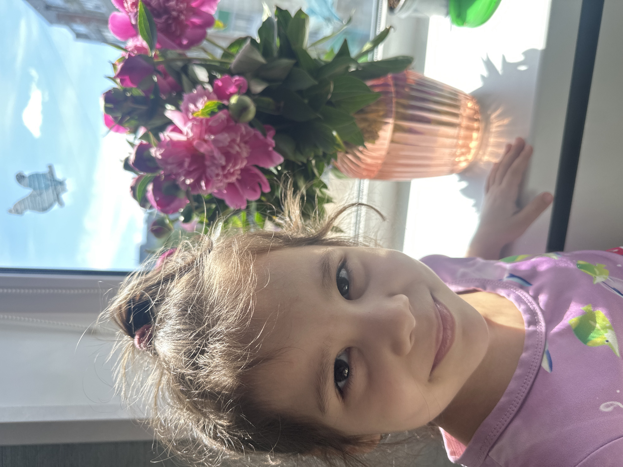 A young child, presumably a girl, with light brown hair styled in a high, messy bun, is positioned in the foreground, facing and smiling directly at the viewer. She has dark eyes and fair skin. Her attire consists of a light purple t-shirt adorned with colorful fish motifs in shades of green, yellow, and white. Her left hand rests on a white windowsill to her right.
On the white windowsill, to the right of the child, a bouquet of pink peonies, some fully open and others still budding, is displayed in a ribbed, translucent, peach-tinted glass vase. Green foliage accompanies the flowers. Sunlight streams through the window, creating pronounced shadows of the vase and flowers on the windowsill, with light refracting through the ribbed glass to form a starburst pattern. A small, indistinct green object is partially visible on the far right of the windowsill.
Beyond the windowsill, a clear blue sky with a single small white cloud is visible through the window. A white rooster-shaped silhouette or sticker is affixed to the upper left pane of the window glass. Distant, out-of-focus buildings are discernible through the window, indicating an urban or suburban environment. The window frame is white. The scene is brightly lit by natural sunlight, suggesting a daytime setting in the city of Unknown, Unknown.
