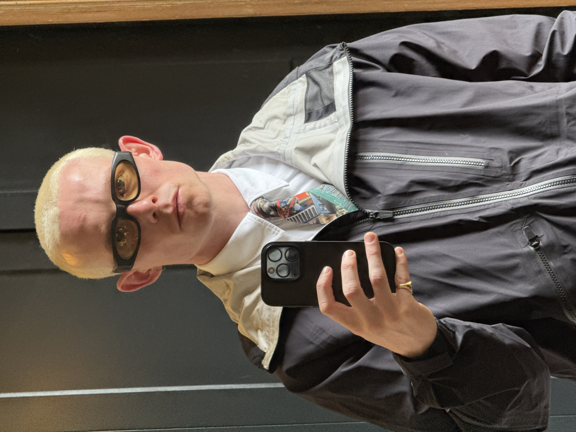 A person with short blonde hair and tinted glasses is holding a black smartphone up to take a selfie. They are wearing a white shirt with a colorful pattern on the collar, underneath a jacket. The jacket is two-toned, with the top part in black and the collar in a beige color. The person is wearing a gold ring on their finger. The background is a gray wall and a wooden beam or trim at the top of the frame. There is no indication of the location being London.