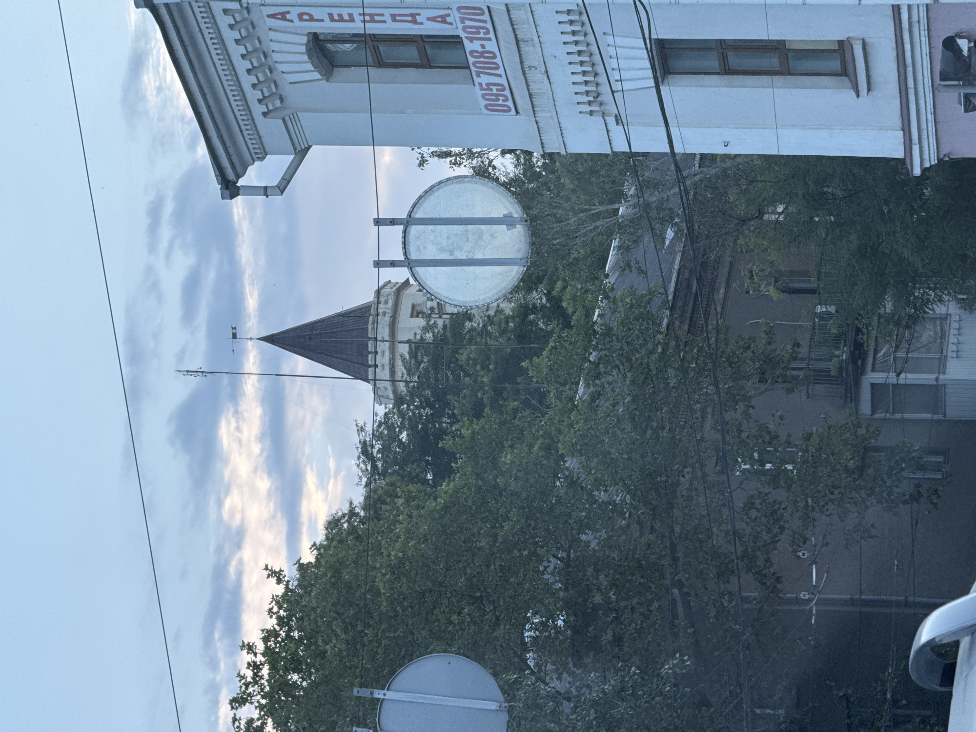 The image shows an overcast sky over a city landscape in Odesa, Ukraine. In the foreground, lush green trees obscure much of the view, with power lines crisscrossing the scene. To the right, a light-colored building with ornate architectural details and a dark-tiled roof is visible. A sign on this building displays the word "АРЕНДА" (rent in Ukrainian) in red capital letters, followed by a phone number "095 708-1970". To the left of this building, a large, circular, unadorned metal sign is suspended by wires. Further in the background, the pointed tower of a building, possibly a historical structure, rises above the trees. Beneath the tower, a series of residential buildings with balconies and windows are dimly visible through the foliage. The overall impression is an urban scene with a blend of old and new architecture, partially hidden by dense greenery.