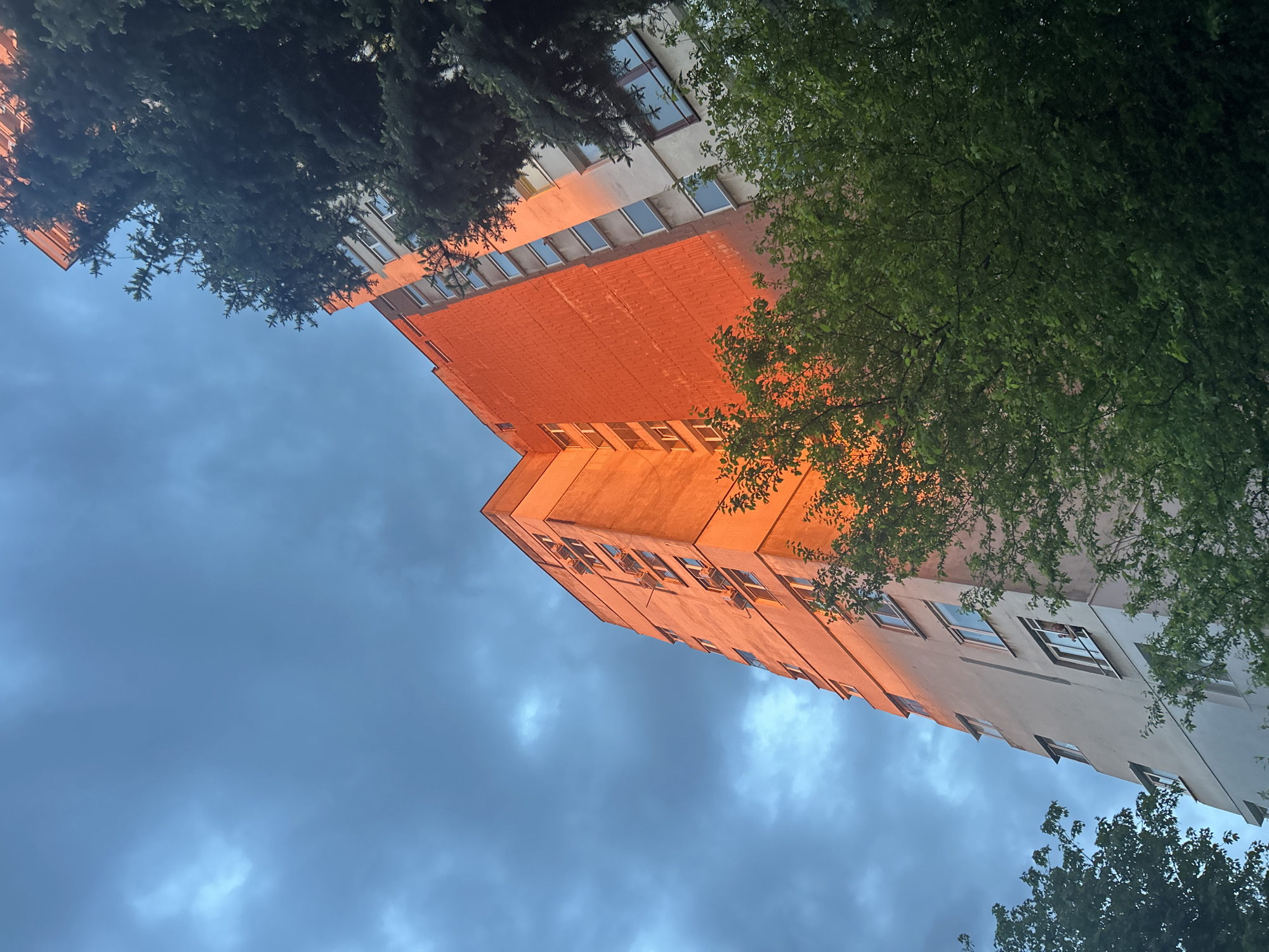 From a low angle, the photo captures a tall building with a mix of orange and white facade, framed by tree foliage. The sky above is blue with some cloudy patches. The location appears to be Sofia, Bulgaria, judging by the architecture style.