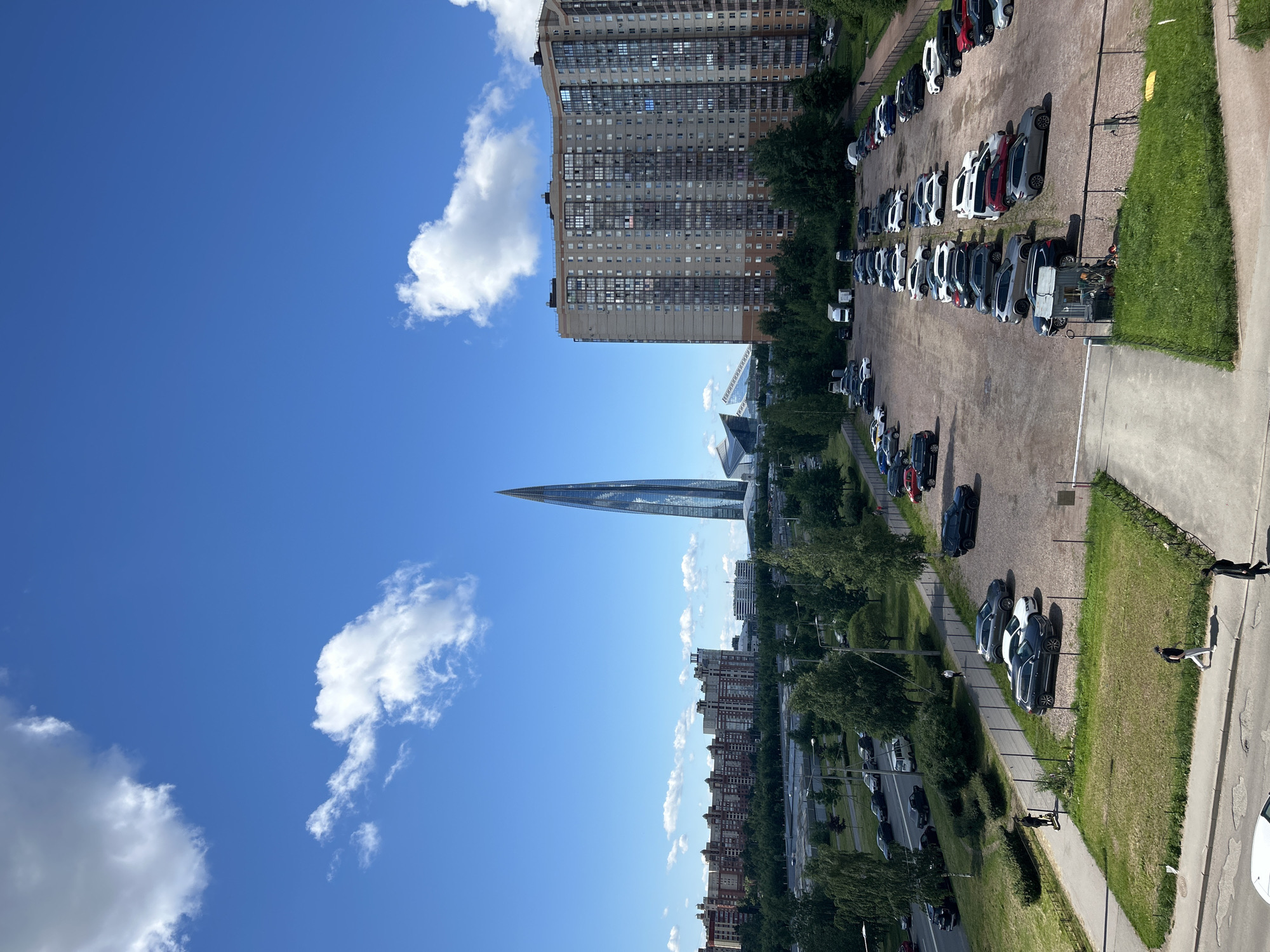 This is an eye-level, high-angle outdoor, daytime shot of the Lakhta Center in Saint Petersburg, Russia. The Lakhta Center is a modern skyscraper with a distinctive twisting glass facade. In the foreground, a large, paved parking lot is filled with numerous parked cars, predominantly SUVs and sedans. A few pedestrians are visible walking on a sidewalk adjacent to the parking lot. In the middle ground, there is a road with cars driving on it, bordered by trees and a green median strip. Behind the road, there are several multi-story residential buildings with many windows. The background features the Lakhta Center prominently, along with other modern architectural structures and a clear blue sky with scattered white clouds. The overall scene depicts an urban environment with a mix of residential buildings, commercial infrastructure, and a notable architectural landmark.