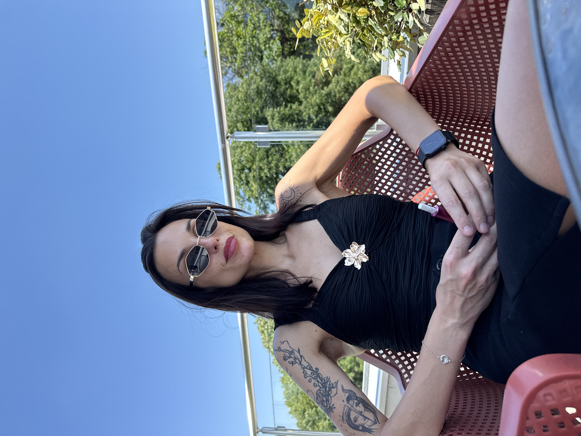 A woman sits in a red, outdoor chair. She is wearing a black dress with a flower brooch, black sunglasses, and a bracelet on her left wrist. She also has a smart watch on her right wrist and tattoos on both arms. The background includes green trees, a blue sky, and a metal railing. The location is likely Pleven, Bulgaria.