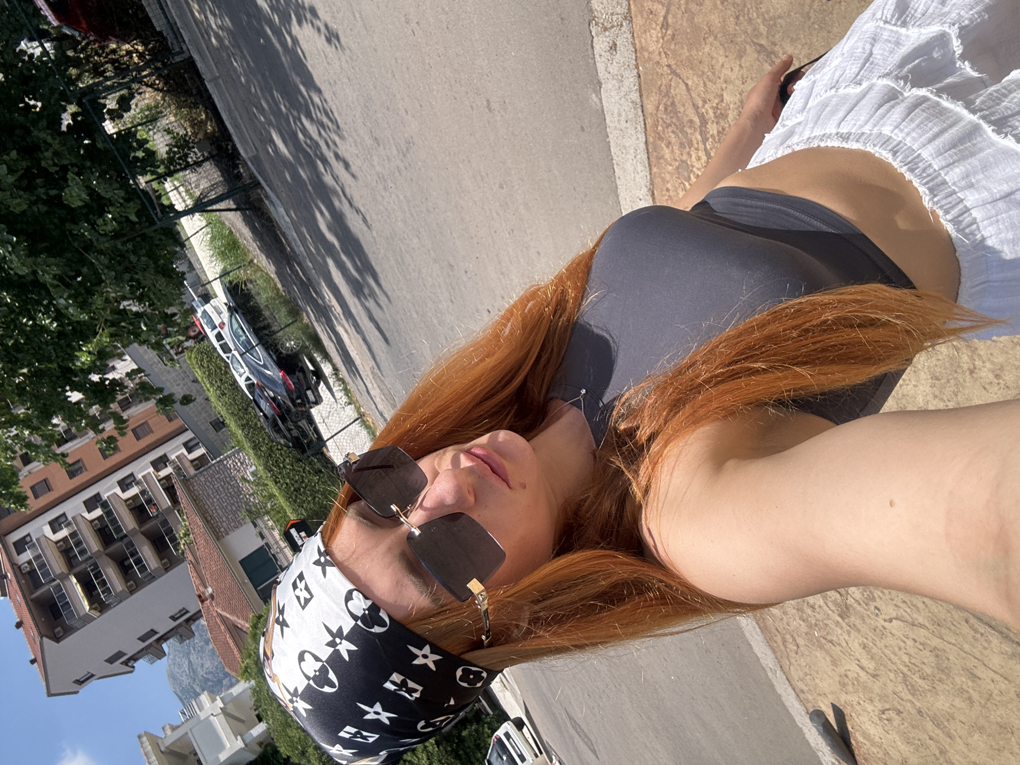 The image depicts a young woman with long, reddish-orange hair and fair skin, wearing a dark grey crop top, a white and black patterned bandana, and oversized sunglasses. She is also wearing a white, ruffled skirt. The woman is holding the camera for a selfie, looking towards the lens.
The background suggests an outdoor urban environment, likely in Budva, Montenegro. A paved road with cars parked along the side is visible. In the distance, there are buildings with multiple stories, characteristic of Mediterranean architecture, and lush green trees. The sunlight casts shadows on the road, indicating it is daytime. The overall impression is of a casual summer day in a coastal city.