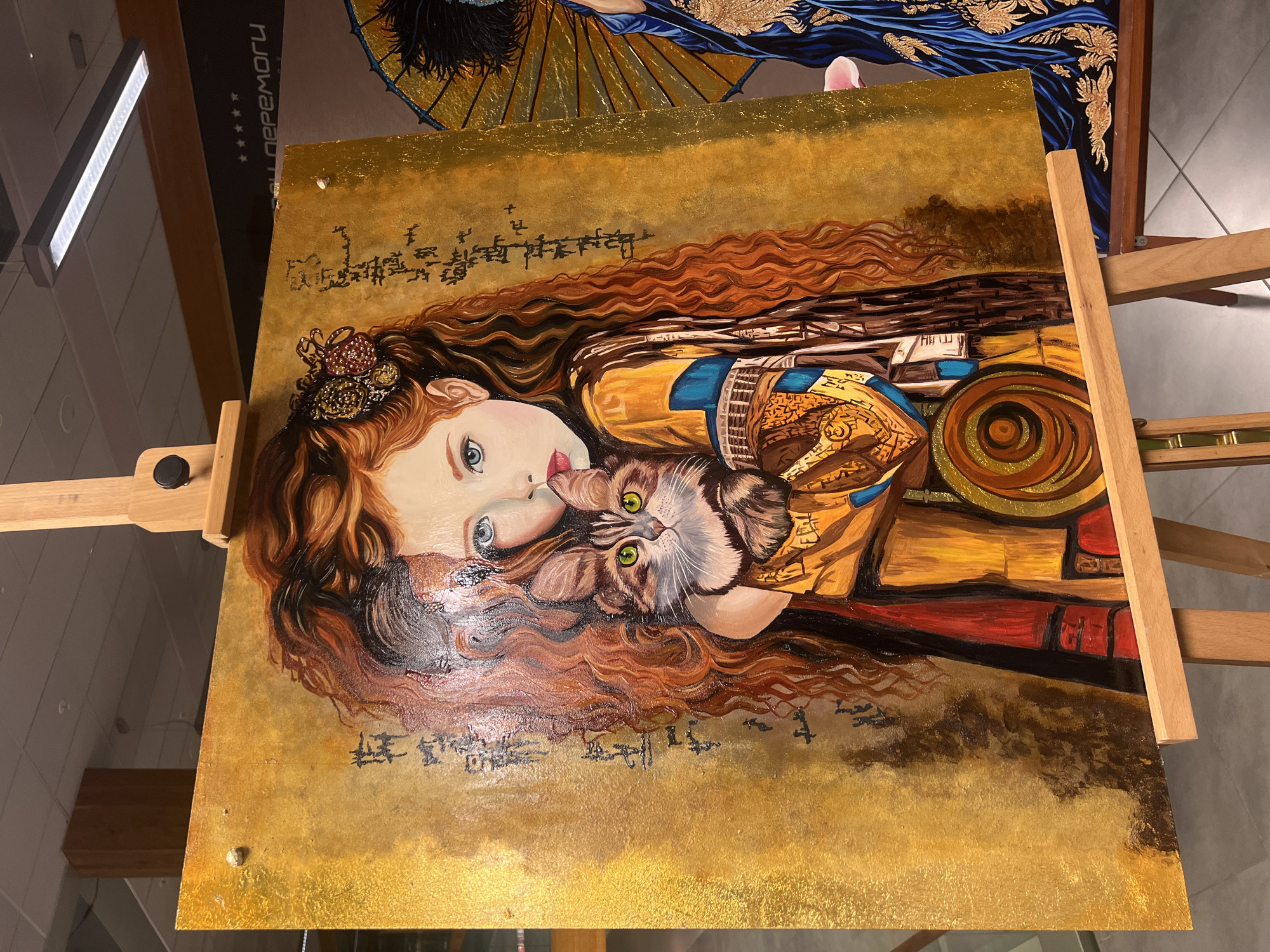 A painting on an easel depicts a young girl with long, wavy red hair holding a tabby cat. The girl has blue eyes, rosy cheeks, and is wearing an ornate yellow and blue outfit with red accents. The background of the painting is a textured gold color with vertical lines of black script. The painting is on display indoors, possibly in a gallery or exhibition space, with a wooden easel supporting it. To the right of the main painting, another artwork featuring a person in a blue, embroidered garment with a golden umbrella is partially visible. The lighting suggests an indoor environment with overhead lights. The floor appears to be tiled.