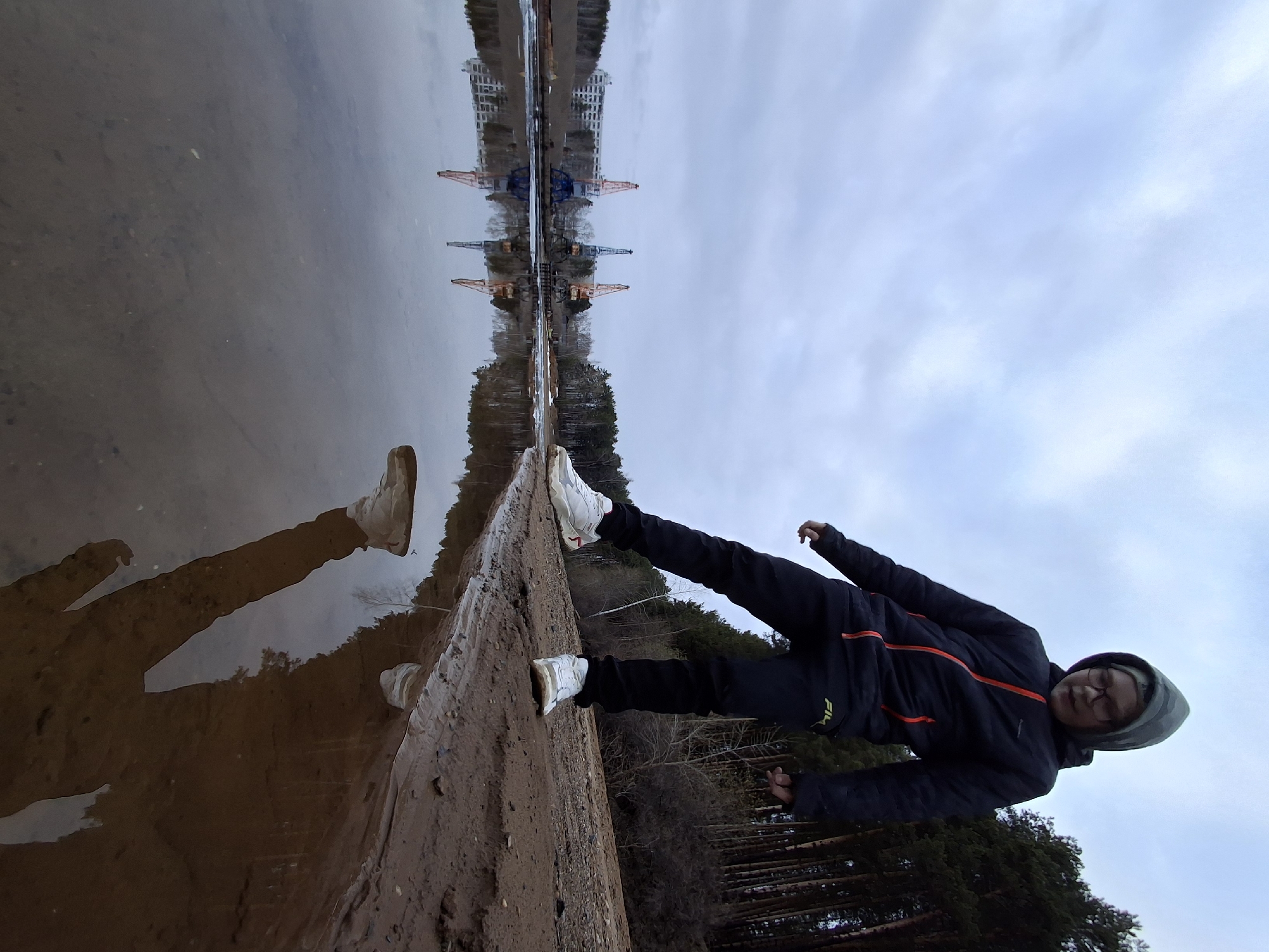 The image, originally rotated 90 degrees clockwise, depicts a young person posing on a muddy bank beside a calm body of water, likely a river or lake, in Chaikovsky, Russia. The person, appearing to be a young male, is dressed in a dark puffer jacket with red or orange striped accents, dark pants, and white athletic sneakers. A grey and black striped hood or beanie covers their head, and they wear glasses. They stand with one leg slightly bent and arms outstretched, creating a dynamic pose.
A clear reflection of the person is visible in the placid, somewhat muddy water below. The background, across the water, features a dense treeline that separates the water from an industrial area. Several prominent industrial cranes, one blue and others reddish-orange, along with distant buildings, are visible above the treeline. These structures are also sharply reflected in the water, creating a strong sense of symmetry. The sky is overcast with light grey clouds, indicating a cloudy daytime setting. The ground directly beneath the person is a mixture of damp earth and mud. The brand name "FILA" is visible in white text on the lower left sleeve of the person's jacket. The scene captures a casual moment where natural elements meet an industrial landscape.