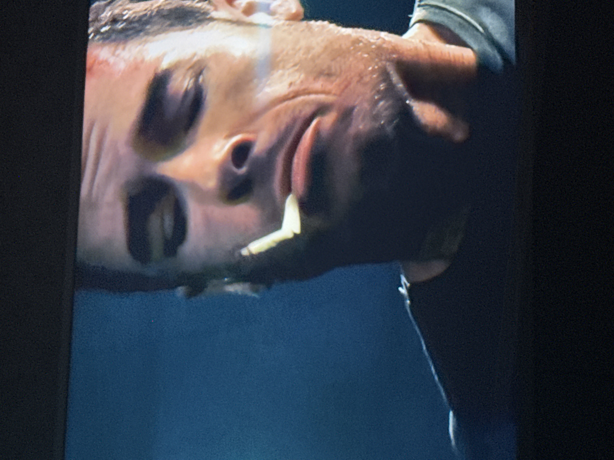 The image shows a close-up of a man's face, appearing to be sweating and possibly underwater or in a humid environment. There is a white object near his mouth that is not fully visible. The background is a blurred, dark blue color. The image is likely a photograph or still from a video. The city of Mytishchi, Russia is not visible in this image.