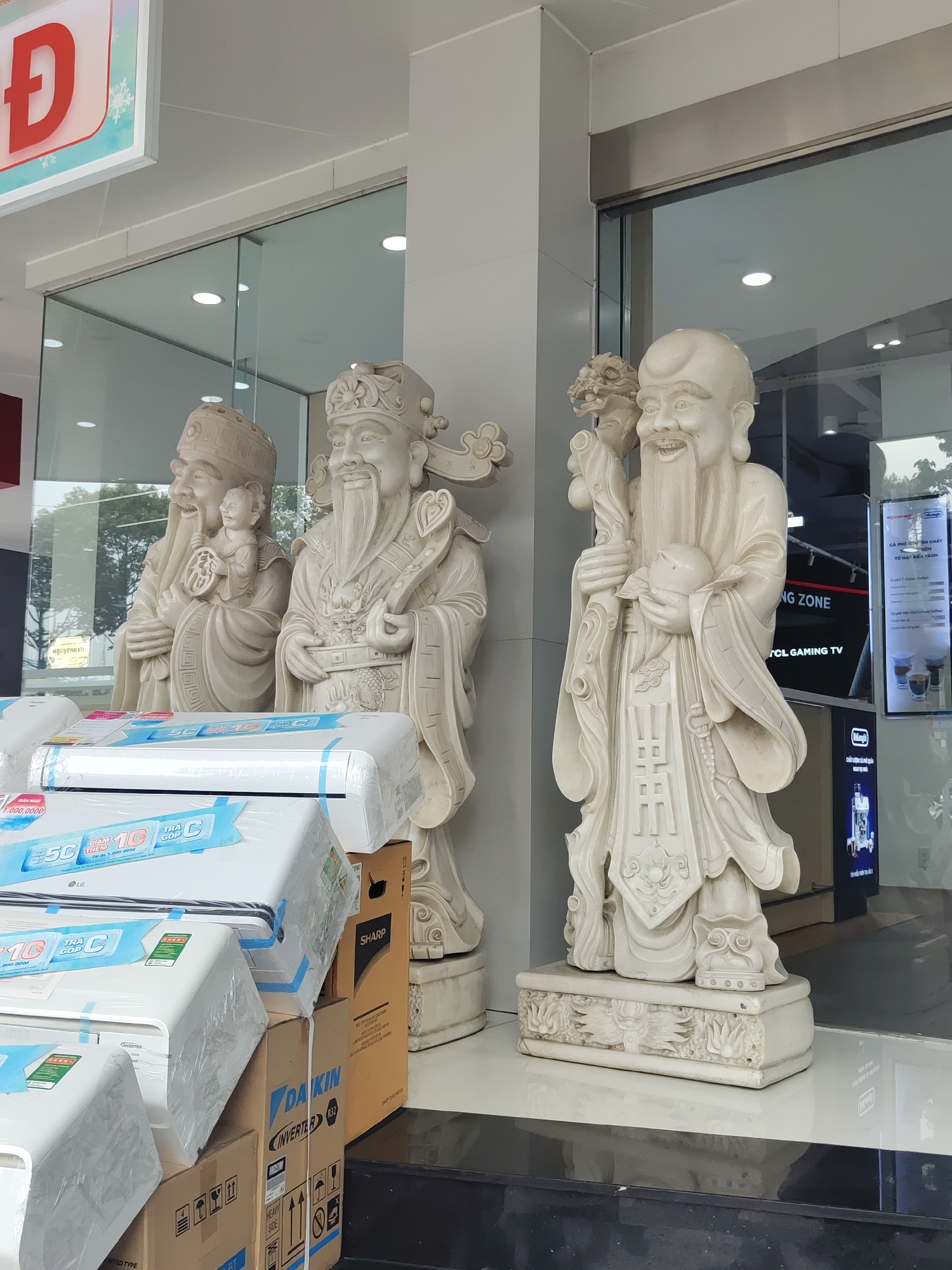 The photograph shows the exterior of a store in Thủ Đức, Vietnam, featuring three white statues of East Asian deities. The statues are standing in front of large glass windows, and in front of two of the statues is a stack of boxed air conditioning units from the brands LG, Daikin, and Sharp. The sign of the store has a red letter “Đ” on it. Through the glass windows, one can see a television displaying “TCL GAMING TV.” A sign inside the store advertises coffee.