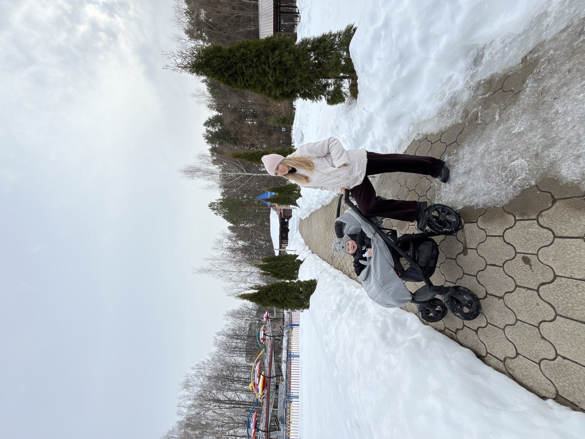 An outdoor winter scene is depicted in the Park Kultury i Otdykha in Chaikovsky, Russia. The image features one adult female and one infant on a paved pathway.
The pathway, constructed from light-colored interlocking concrete pavers with a wavy pattern, extends from the bottom-center towards the upper-left. Significant snow cover is present on either side of the pathway, forming distinct banks. Sections of the pathway near the adult's feet display visible ice and slush.
On the right side of the pathway, an adult female stands, facing left with her gaze directed towards the mid-right. She has blonde hair and is dressed in a light pink knit beanie, a light beige or off-white quilted winter jacket, dark-colored pants, and dark shoes. Her left hand rests on the frame of a stroller.
To her left, an infant is seated within a grey and black four-wheeled stroller, identified by the visible "PRAMY" branding. The infant wears a grey hat with ear-like embellishments and a dark jacket, and is observed smiling towards the camera. A small white item is present in the stroller's seat.
The background on the left side of the scene reveals an amusement park area, separated by a low, colorful metal fence comprising red, blue, yellow, and green vertical bars. Several stationary, brightly colored 'flying saucer' type amusement rides, featuring red, yellow, blue, and green gondolas, are visible beyond the fence. The landscape includes numerous bare deciduous trees and dark green coniferous trees, forming a dense line in the distant background. A small brown-roofed building and a smaller structure with a blue roof are discernible in the mid-ground. A series of conical evergreen trees are aligned along the pathway on the right side. The sky is uniformly overcast and light grey.