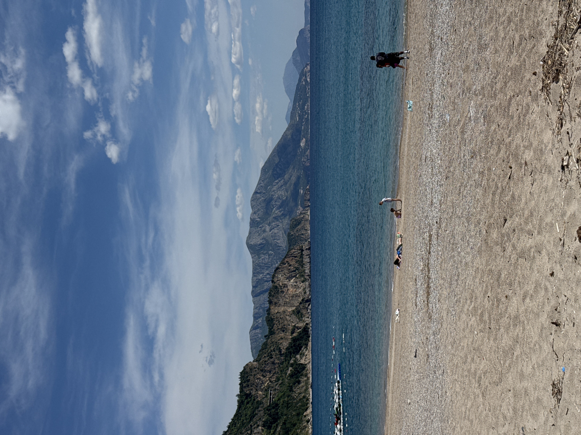 The image depicts a beach scene in Prijevor, Montenegro, with mountains in the background. The sky is blue with scattered white clouds. The beach is sandy with some pebbles. Several people are present: two individuals walking on the beach, two more appear to be standing in the water, and two are lying on the sand. Debris such as sticks and litter is on the beach. In the water, a series of white floats are visible.