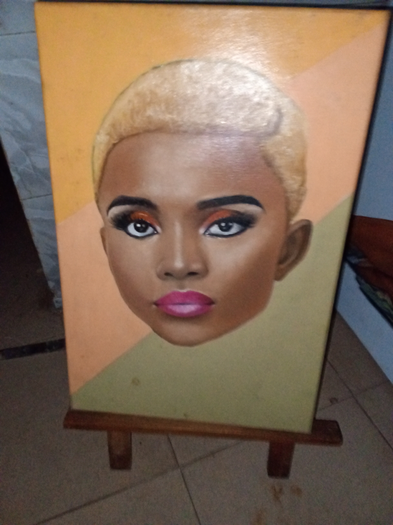 The image displays a vibrant portrait painting on a canvas, presented on a wooden easel in an indoor setting in Ado, Nigeria. The painting is a close-up of a woman's head, featuring dark skin and short, blonde hair. Her facial features are highlighted with striking makeup: dark, defined eyebrows, elaborate eye makeup including bright orange eyeshadow, black winged eyeliner, and long, dark eyelashes. Her dark eyes have distinct white sclera, and her full lips are painted with a bold, bright pink lipstick. The woman's expression appears neutral or serene.
The background of the painting is abstract, divided diagonally into two color blocks: a warm orange-yellow in the upper left section and a muted green-khaki in the lower right section. The canvas itself is rectangular and appears to be made of a textured material, possibly canvas. It is supported by a simple wooden easel with visible legs. The floor beneath the easel is covered with light-toned tiled flooring, suggesting an indoor environment. There is no specific activity taking place other than the display of the artwork. The time of day is not discernible from the indoor lighting. No text is visible in the image.