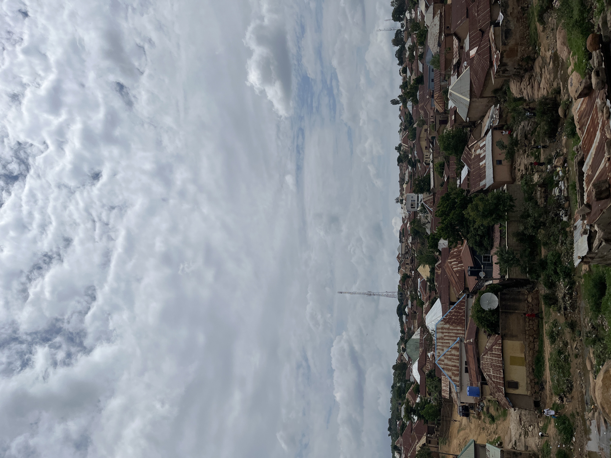 This image captures an elevated view of a densely populated residential area in Jos, Nigeria, under an overcast sky. The upper two-thirds of the frame are filled with a thick, textured canopy of grey and white clouds, with intermittent glimpses of pale blue sky visible through breaks.
The lower third of the image depicts numerous single-story and a few two-story buildings, characteristic of a high-density urban or peri-urban settlement. The structures feature predominantly rusted corrugated metal roofs, displaying various shades of orange and brown, interspersed with some newer or painted roofs in blue, green, and unrusted metallic tones. The walls of the buildings are generally constructed from earth-toned materials.
A tall, multi-tiered communication mast, likely a cellular tower, stands prominently on the left side of the midground, extending well above the rooftops. In the lower-center, a white, circular satellite dish is affixed to the exterior wall of a building. Blue and black cylindrical water storage tanks are visible on elevated stands adjacent to some houses.
The terrain is uneven and rocky, with unpaved dirt paths and tracks winding through the settlement, exhibiting signs of erosion and muddy patches. Sparse green vegetation, including small trees and bushes, is scattered among the buildings and along the pathways.
Several individuals are present in the lower foreground and midground. On the far left, two figures, one in a white top and another stooping, are near a muddy depression or small stream. A lone figure in a red top walks along a dirt path towards the right in the lower-center. A small cluster of three to four individuals is observed near rocks and vegetation in the lower-right midground. The foreground itself consists of rugged, rocky ground with visible detritus.