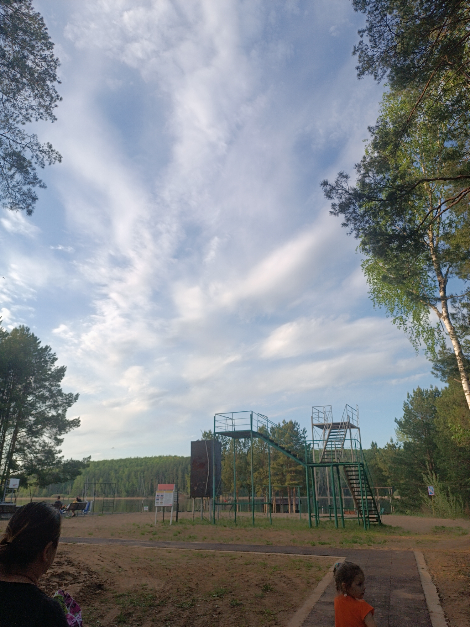 A wide shot captures a natural scene. In the foreground, a person stands to the left, their back to the viewer, while a child in an orange shirt looks away. In the midground, there is a two-level metal structure with stairs, resembling a diving platform, in the center of the image. A lake is behind a perimeter fence, beyond which stands a dense forest. The sky is filled with wispy clouds. Trees frame the image on either side. The location is Unknown, Unknown.
