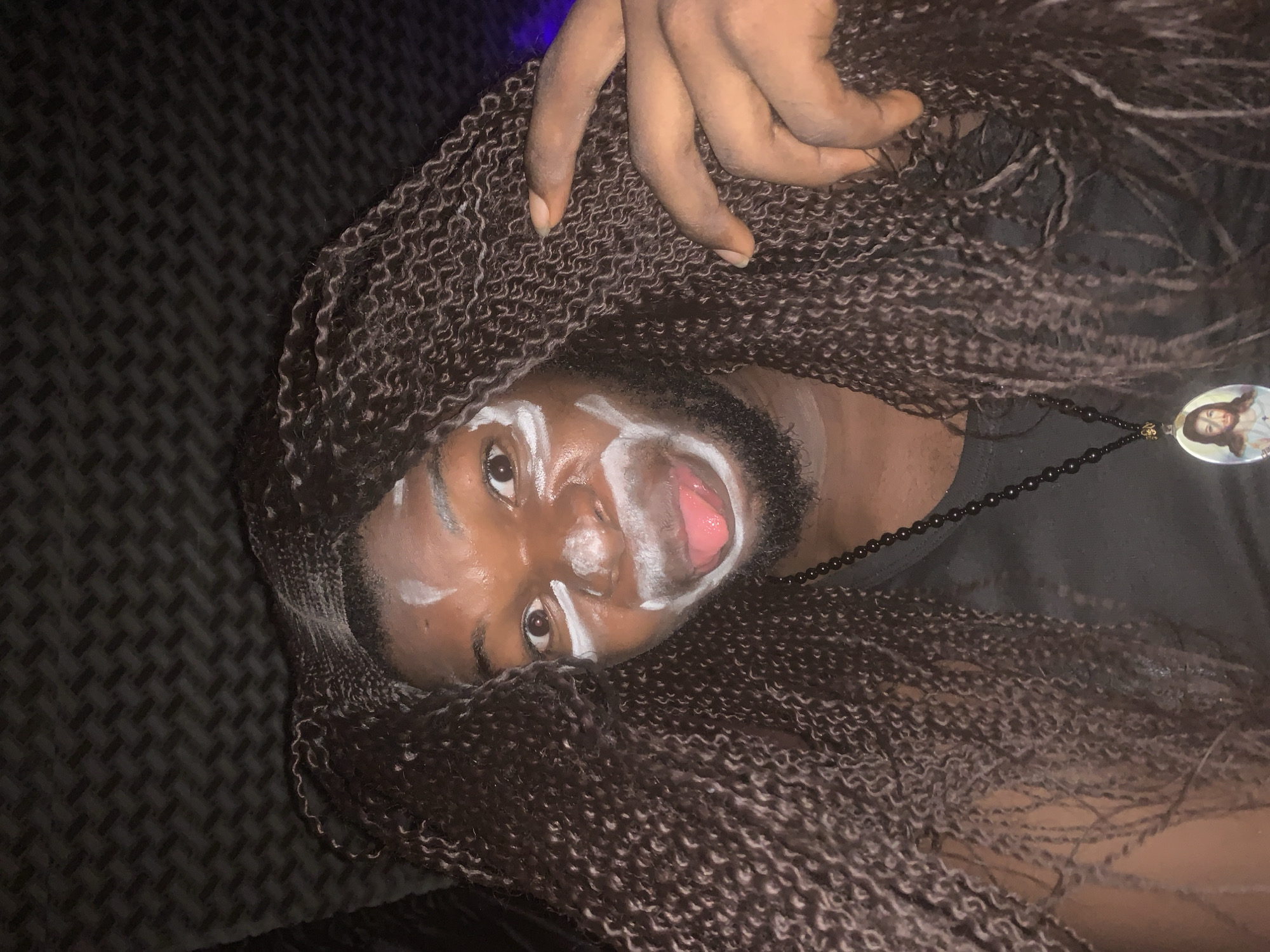 The image presents a person with dark skin, a beard, and long, dark, intricately braided hair. Their face is adorned with striking white markings applied around the eyes, nose, forehead, and cheeks. The person looks directly at the viewer with an open mouth and their tongue slightly extended.
A hand with dark skin is visible at the top of the frame, appearing to interact with or arrange the voluminous braided hair. The person is wearing a dark-colored top and a black beaded necklace, from which hangs a circular pendant depicting a classical portrayal of Jesus Christ.
The background is a dark, patterned surface, possibly a woven mat or fabric with a diagonal grid texture, suggesting an indoor environment with low lighting. The image itself is oriented ninety degrees clockwise. The white facial paint, the direct gaze, and the extended tongue could indicate a performance, cultural ritual, or artistic expression. No text is visible in the media file.