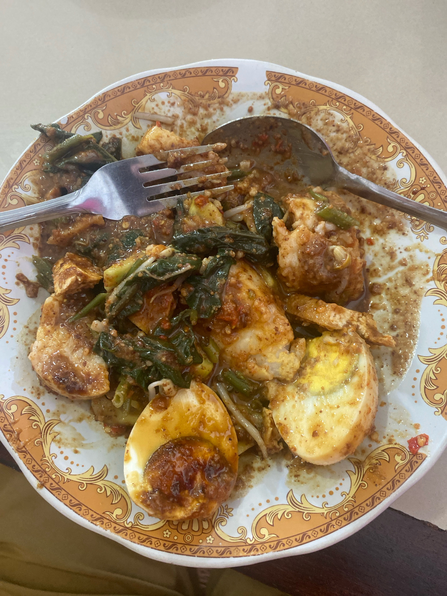 A close-up, top-down shot shows a plate of food on a table, with a fork and spoon resting on it. The dish, which appears to be a type of salad or vegetable stir-fry, is covered in a brown sauce and contains pieces of what look like tofu, vegetables, and hard-boiled eggs. The plate has an ornate orange and white pattern along its rim. The background is blurred and appears to be a light-colored surface, possibly a table or countertop. The overall scene is domestic or casual dining. There is no indication of the location in Labuha, Indonesia.