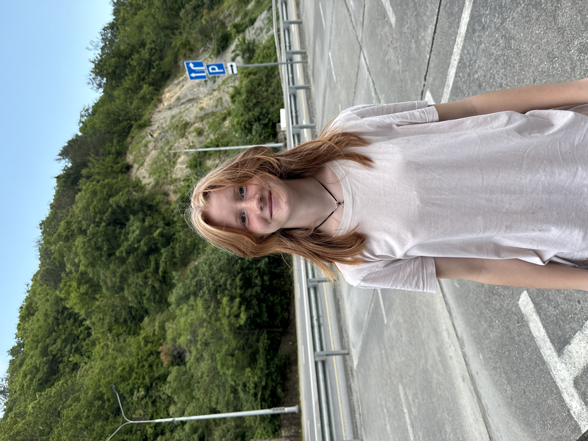 A young female with auburn hair stands facing forward, smiling slightly. She is wearing a light-colored t-shirt and a dark necklace. The background consists of a steep, wooded hillside with a clear blue sky above. In the middle ground, there is a road with guardrails and a blue traffic sign with white symbols indicating a turn and parking. The ground in the foreground is paved asphalt with white line markings, possibly a parking area. The overall setting appears to be a roadside in a mountainous or hilly area. Based on the provided information, this location is in the city of Nebug, Russia.