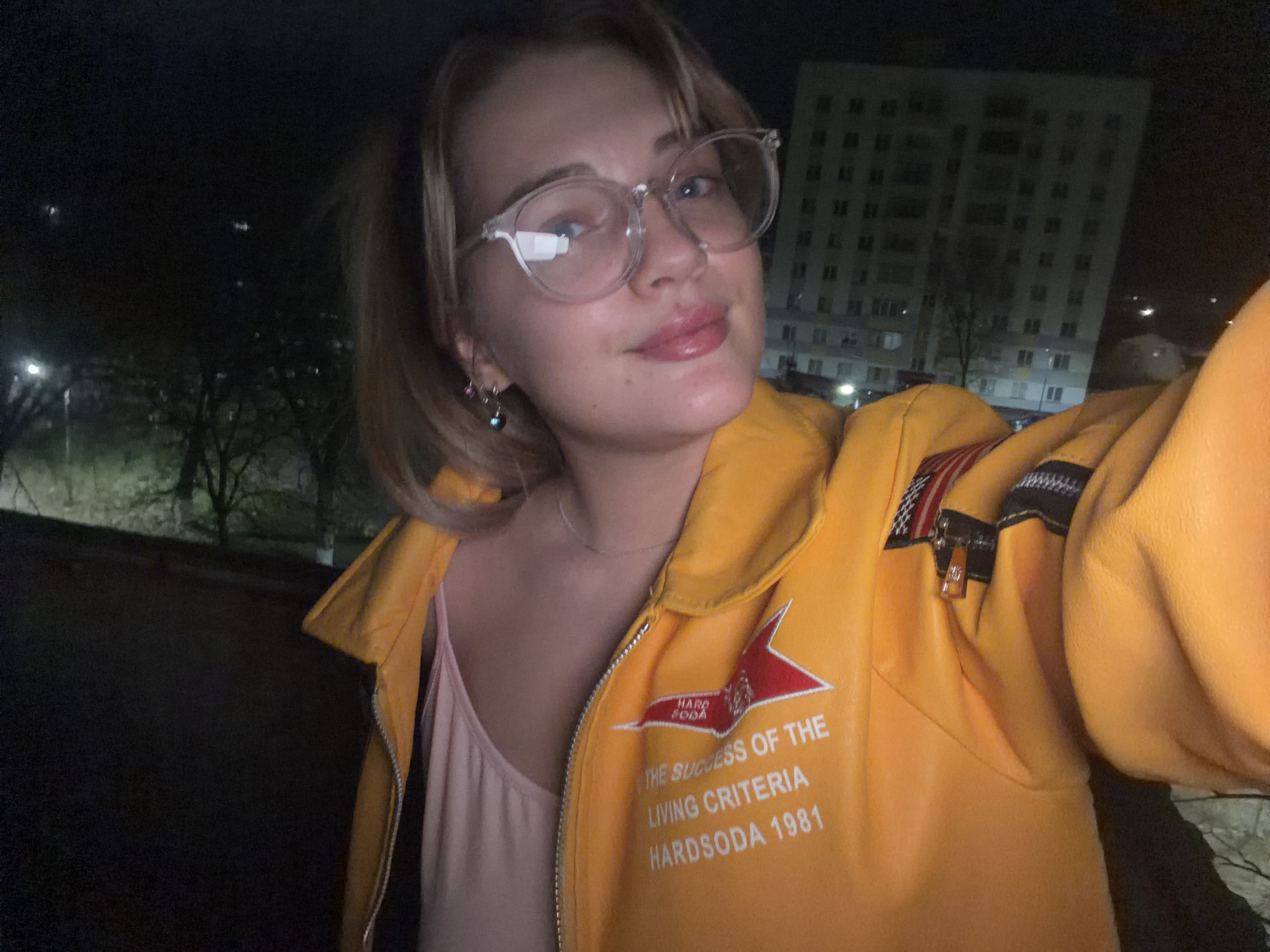 A blonde woman with glasses is posing for a selfie at night. Behind her is an apartment building. The woman is wearing a yellow jacket that says "HARD SODA THE SUCCESS OF THE LIVING CRITERIA HARDSODA 1981". The location is likely in Naberezhnye Chelny, Russia, given the style of the apartment building and the woman's jacket.