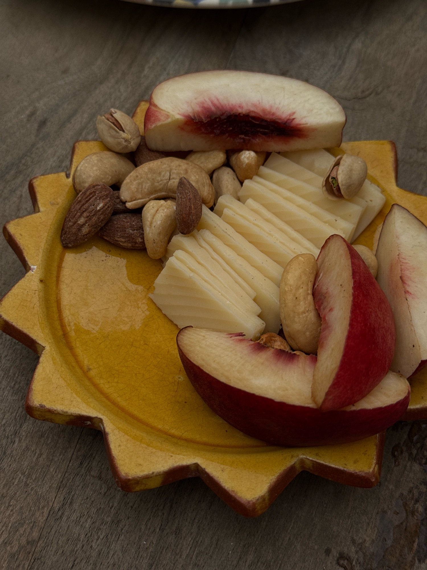 The image shows a close-up, high-angle shot of a yellow, star-shaped dish filled with sliced peaches, cheese, and assorted nuts. The peaches are ripe, with pink and red skin and white flesh, sliced into wedges. The cheese is sliced thinly and arranged in overlapping rows. The nuts include almonds, cashews, and pistachios. The dish is placed on a textured, gray wooden surface, possibly a table. The lighting is natural, creating soft shadows. The scene appears to be a snack or appetizer setting. The image does not contain any explicit information about the city of Essaouira, Morocco, or any identifiable landmarks that would place it there.