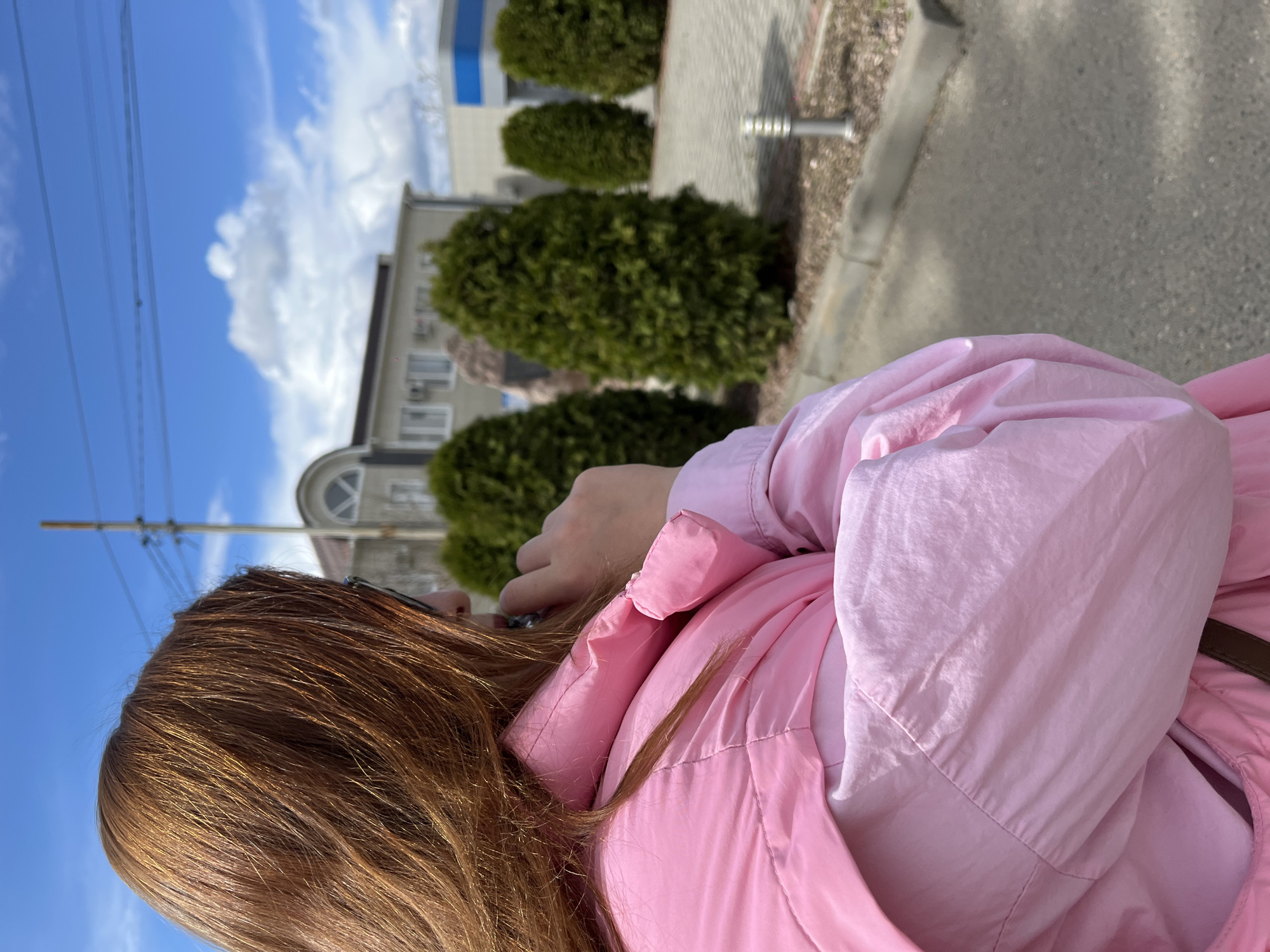 A woman with reddish-brown hair and a pink jacket is on a sidewalk, with a building and greenery behind her. The sky is blue with some clouds. The location appears to be Novopokrovskaya, Russia, given the context.