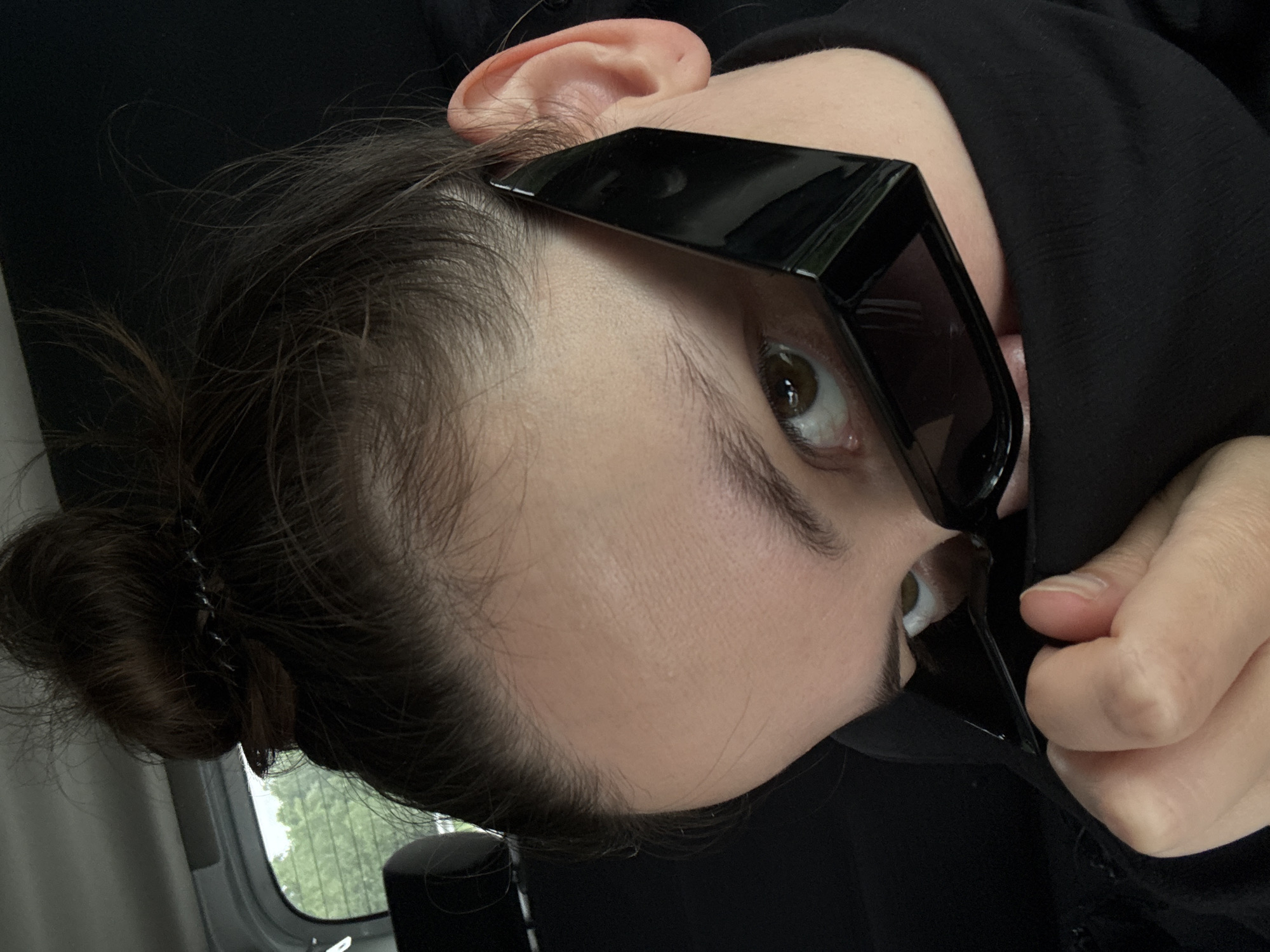 A person with fair skin and dark brown hair pulled into a bun is shown in a close-up, tilted portrait orientation. They are wearing black sunglasses. Only one eye is fully visible, and it is brown. The person is wearing a black garment. Part of their ear is visible. A hand is holding the sunglasses. A window is visible in the background with green trees. The location is not discernible from this image.