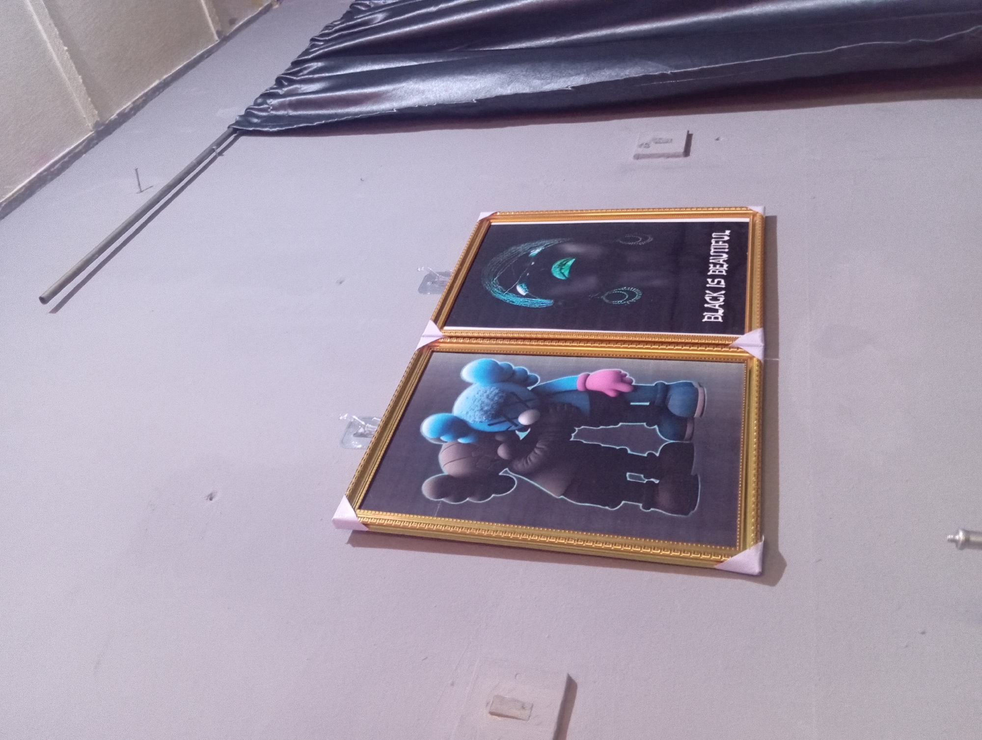 A medium shot captures a wall with two framed pictures hanging on it. The left picture features two cartoon characters in a hug. One character is brown and the other is blue. The blue character is wearing a pink glove. The right picture is a portrait of a person with bright green lips and eyes, outlined with neon blue. The text "BLACK IS BEAUTIFUL" is written at the bottom of this frame. The frames are gold with white corner protectors. To the right of the frames is a dark grey curtain hanging from a metal rod. The wall is painted a light grey color. There is a light switch cover to the left of the pictures.