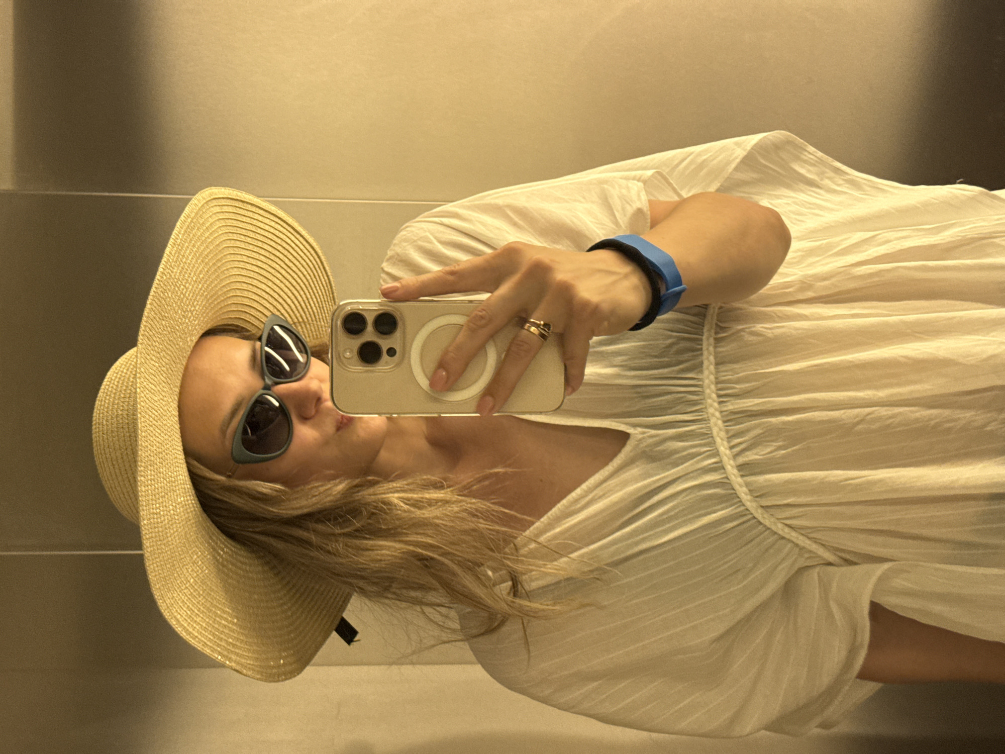 A woman is taking a selfie in what appears to be an elevator. She is wearing a wide-brimmed straw hat, sunglasses, and a loose white dress. She is holding a smartphone in her right hand, which is visible in the frame. A blue wristband is on her left wrist. The background is a plain, reflective surface, likely the wall of an elevator. The lighting is warm and slightly diffused, creating a golden hue. The image quality is good, with clear focus on the subject. The location in Nebug, Russia, is not explicitly indicated in the image itself.