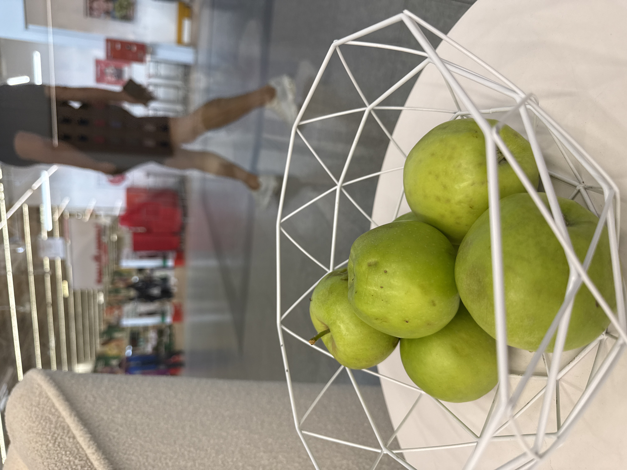 A white wire basket filled with green apples sits on a white table. In the background, blurred by reflections in a glass partition, a person walks through a brightly lit indoor space, possibly a store. The person is wearing a dark top and a patterned skirt, and is carrying a phone in their right hand. The floor appears to be a polished concrete or similar material. The scene is indoors, with fluorescent lighting visible on the ceiling. The location is not specified as Nizhny Novgorod, Russia.