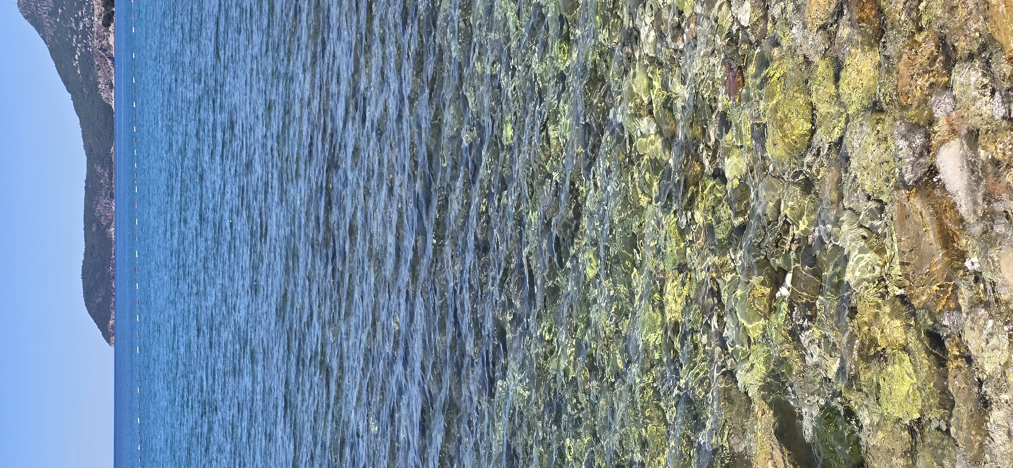 The image depicts a coastal scene in Bar, Montenegro. On the left, calm, clear blue water meets the shoreline where the ocean bed is visible. As the water gets shallower, the bottom of the ocean becomes more visible, revealing a variety of pebbles and stones in shades of grey, brown, and green. The shoreline itself is composed of rocks, stones, and sand. In the distance on the left, a rugged coastline is faintly visible against the blue sky.