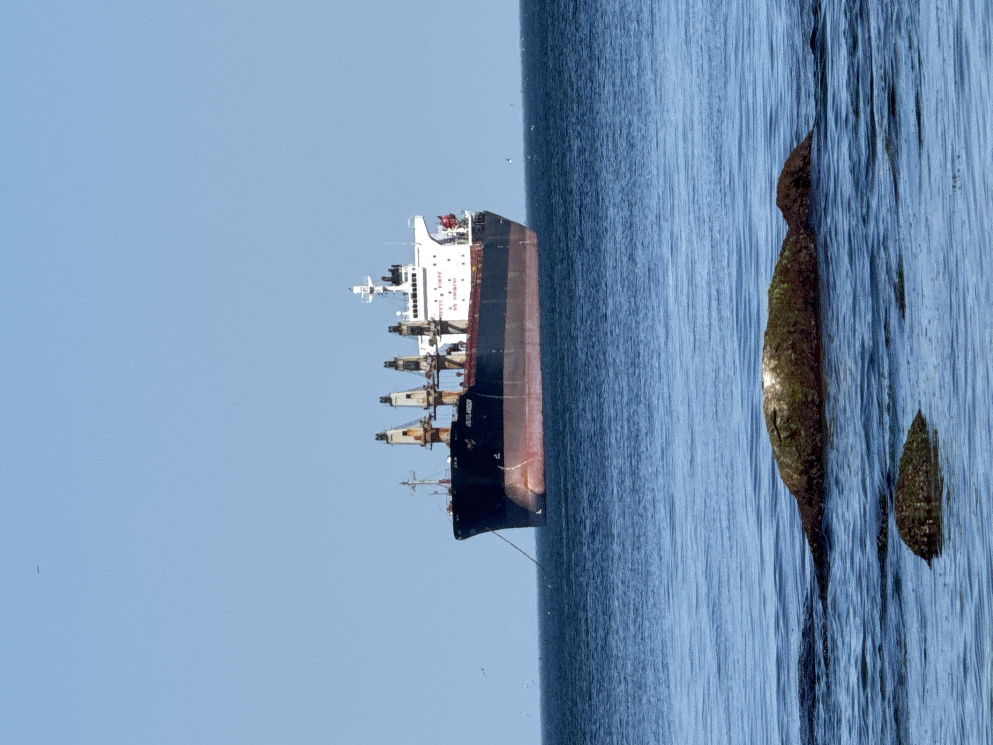 The image presents an inverted view featuring a large cargo ship on the left side, set against a clear blue sky. The ship is a multi-decked vessel, equipped with cranes and navigational equipment. The hull is painted black and red, and its name, "OUTLANDER", is visible in white lettering.
On the right, there is water, and two rocks submerged in the water. It's hard to determine the specific location in Varna, Bulgaria without more contextual information, however it shows the coastline with a vessel moored offshore.