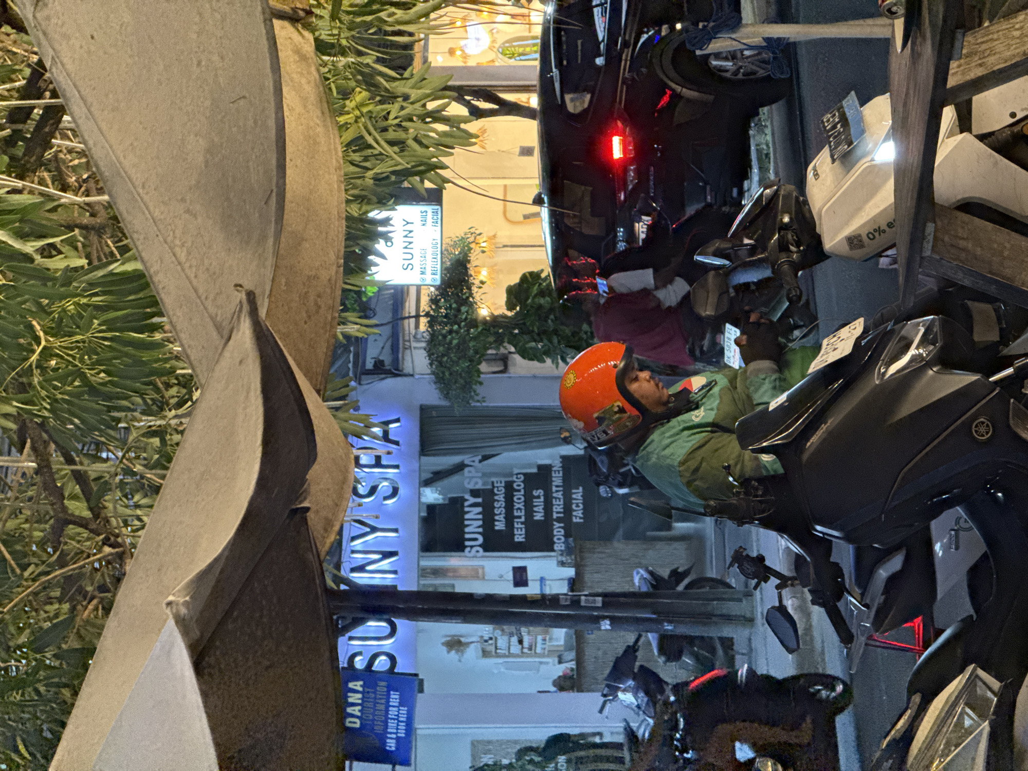 In North Kuta, Indonesia, at night, a street scene unfolds under the canopy of an outdoor seating area, likely at a spa. A person wearing an orange helmet and a green jacket is seated on a motorcycle, looking down at a mobile phone in their hands. The motorcycle is parked in front of a storefront, "SUNNY SPA," which is illuminated with blue neon lights. Another sign, also for "SUNNY SPA," lists services such as massage, reflexology, nails, body treatments, and facial. The storefront displays internal lighting and appears to be a commercial establishment. Parked motorcycles and a black car are visible in the background, with some of the motorcycles displaying license plates. Trees and foliage are present, suggesting an outdoor or semi-outdoor environment. The overall lighting indicates it is nighttime.