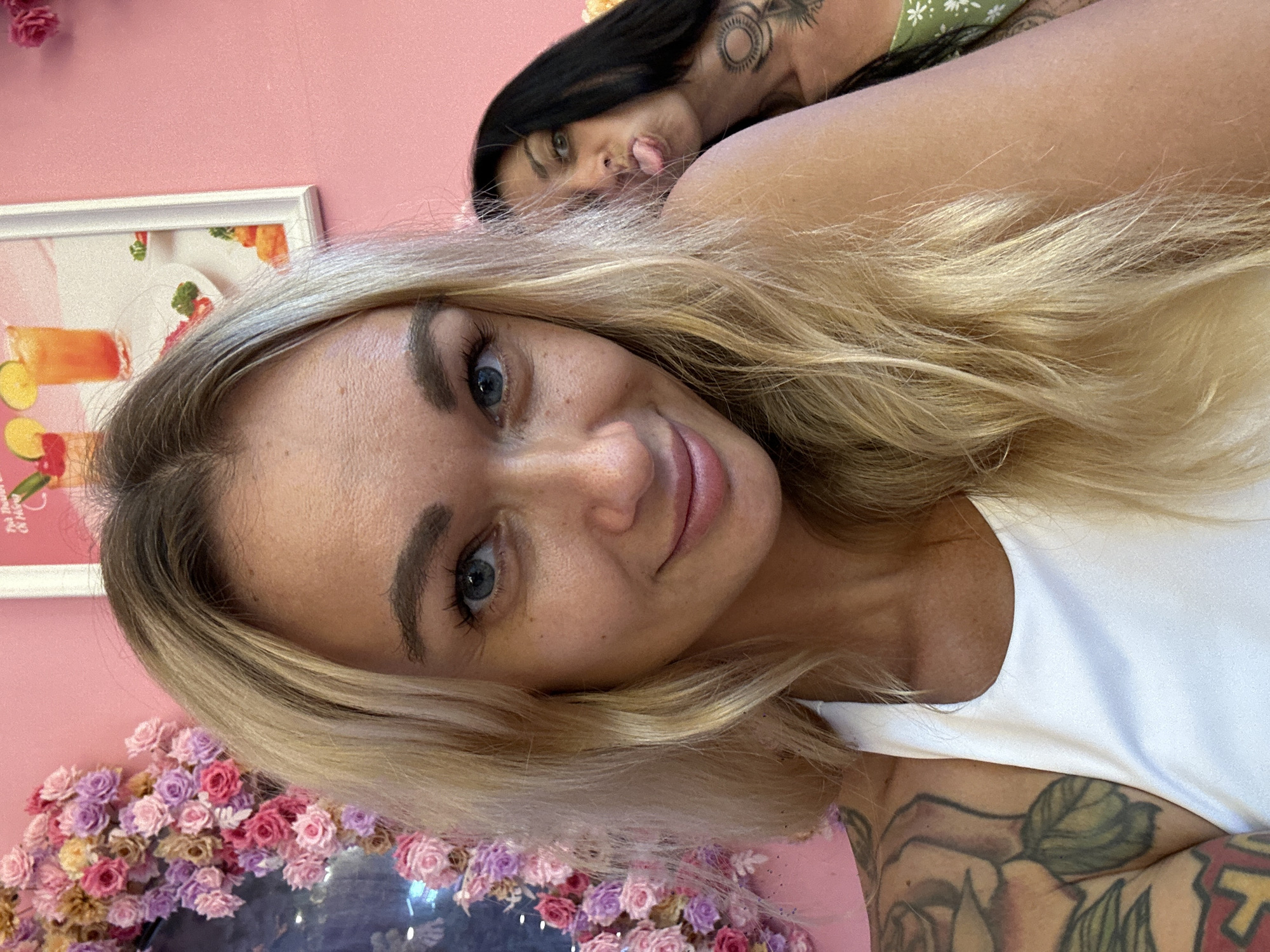 Two women are featured in a close-up, slightly tilted photo taken indoors, likely a selfie. The dominant subject in the foreground is a woman with long, wavy blonde hair, blue eyes, and fair skin, looking directly at the camera with a soft, friendly smile. She has light freckles and noticeable eyebrows. She wears a white sleeveless top, and a large, colorful floral tattoo with green leaves is visible on her right upper arm.
Behind her, partially obscured and positioned higher in the frame, is another woman with long, dark hair. She is looking towards the camera and playfully sticking out her tongue. A prominent tattoo with circular and tribal-like designs is visible on her shoulder/neck area, and she appears to be wearing a green top with a white floral pattern.
The background consists of a solid pink wall. On the left side, a white-framed picture hangs, depicting vibrant illustrations of what appear to be fruity drinks or cocktails with various garnishes. Faint, blurry text on the picture suggests "The Dessert" and possibly "Pina" and "CA". Below the foreground woman, a cluster of small, artificial roses in shades of pink, purple, and yellow creates a decorative border, possibly adorning a mirror or table. The overall mood of the image is lighthearted and cheerful, set in a brightly lit, aesthetically pleasing environment.