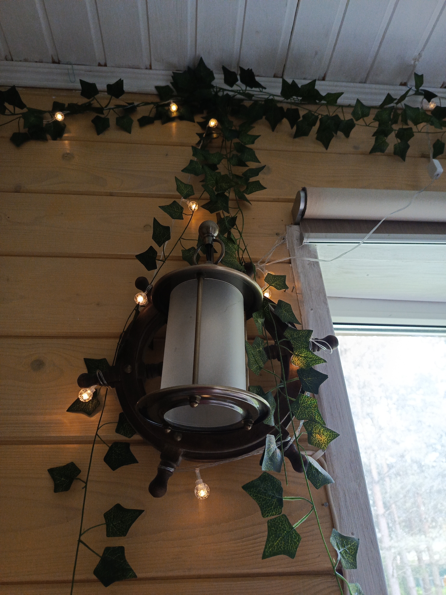 A ship's wheel wall lamp hangs on a light brown wood-paneled wall in a room in Северный, Russia. The lamp is encircled with faux ivy garland and small, round fairy lights. A window with a white frame and white roller shade is to the right of the lamp, revealing a view of trees. The wall above the lamp also has faux ivy and fairy lights.
