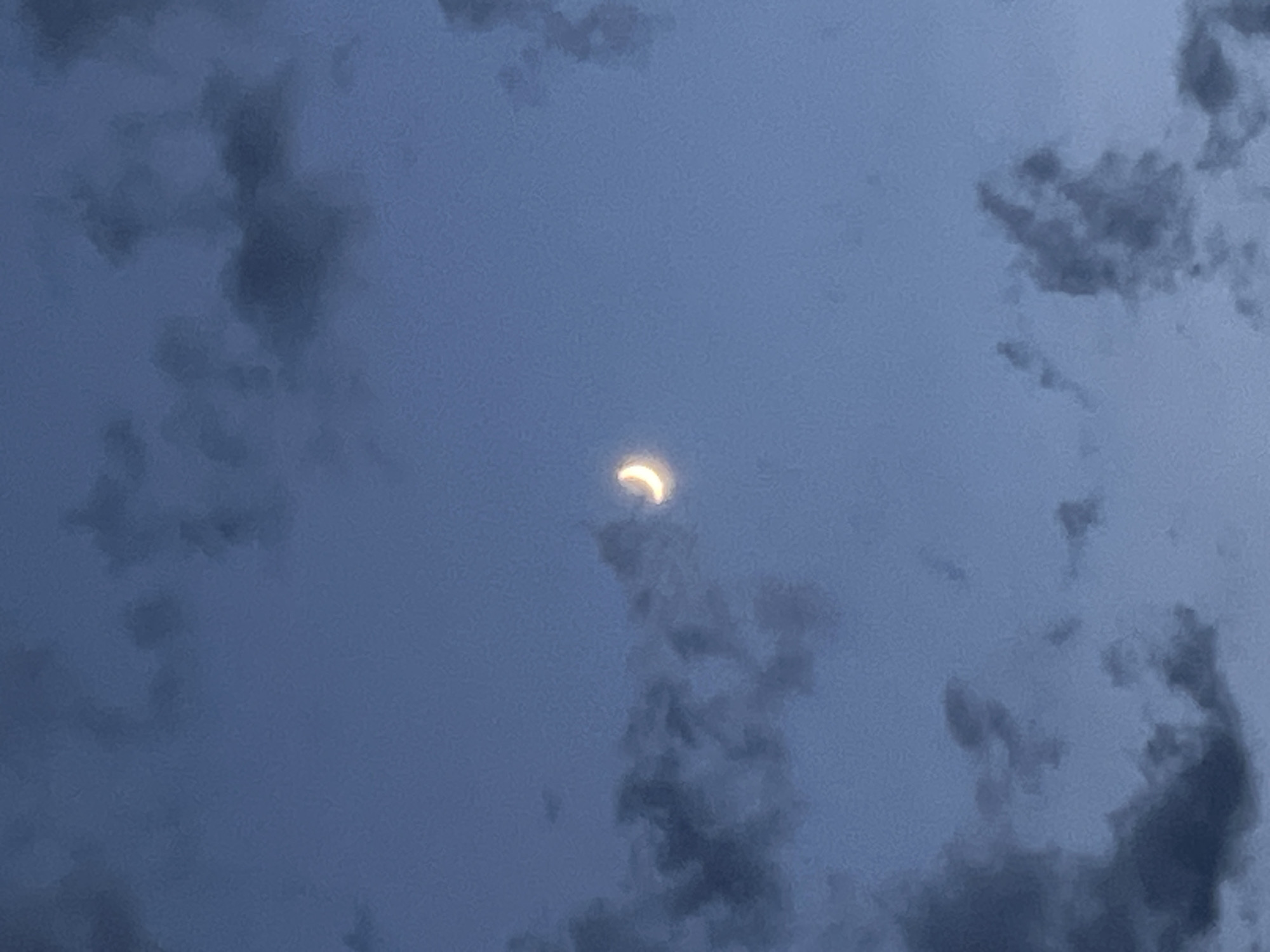 The image depicts a crescent-shaped celestial body, likely the Moon or a solar eclipse, visible in a cloudy sky. The sky is a muted blue-gray, indicating either dawn, dusk, or an overcast day. Darker, amorphous clouds are scattered across the frame, partially obscuring the sky. The celestial body, appearing as a thin sliver of light with a yellow-orange hue, is positioned slightly left of center, nestled amongst the clouds. Its luminous crescent shape stands out against the darker background. The overall mood of the image is somewhat ethereal and mysterious, with the clouds adding a sense of depth and texture to the scene. There is no visible text in the image. The location context is Sochi, Russia, but there are no specific visual cues within the image that definitively point to this location. The scene appears to be an outdoor setting, likely viewed from ground level, observing the sky. The activity is the observation of a celestial event through a partly cloudy sky.