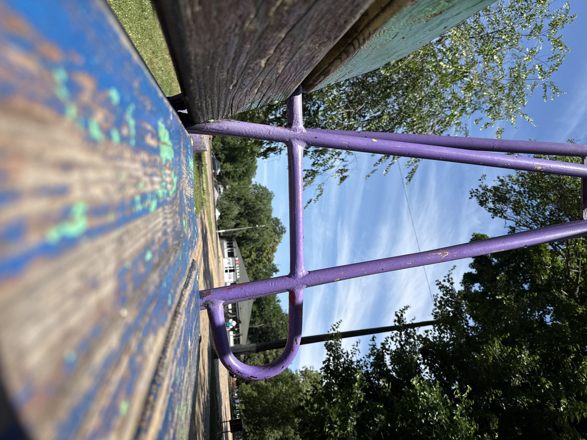 An outdoor shot captures a close-up perspective of a weathered, blue and green painted wooden bench. The bench is positioned low to the ground, with the front edge in sharp focus, showing peeling paint and wood grain. Behind the bench, a purple metal swing set frame is visible, its tubes showing signs of wear and tear with flecks of yellow paint. In the background, a paved area leads to a building with a green roof, a sign displaying Cyrillic text that reads "Я КЕБАБ" (Ya Kebab), indicating a food establishment. Several trees with lush green foliage surround the area, and the sky is clear blue with scattered wispy clouds. A dark-colored car is parked in the distance. Two indistinct figures are walking in the background near the building. The scene appears to be set in a public park or recreational area within the city of Piatykhatky, Ukraine, under bright daylight conditions.