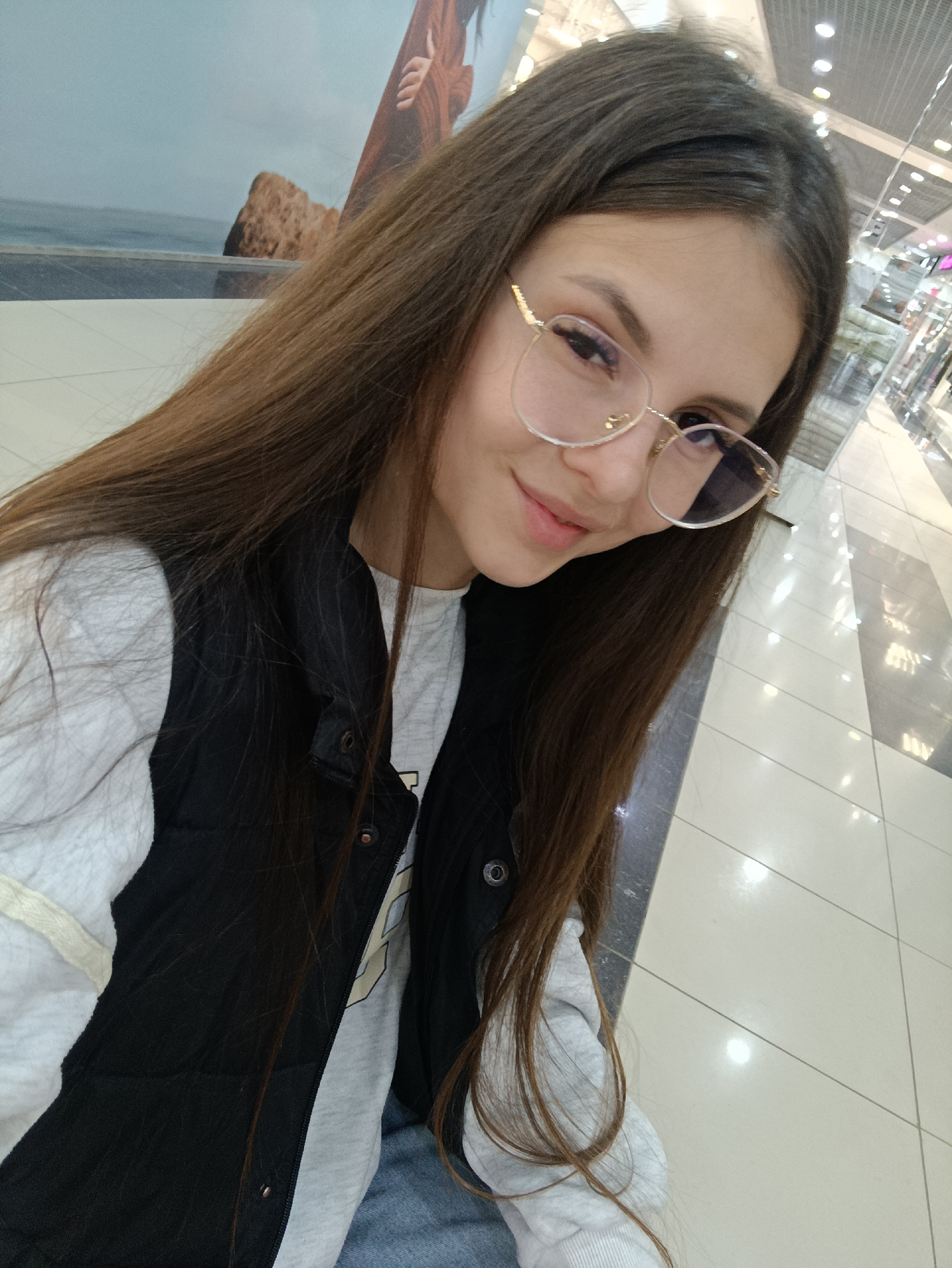 A young woman with long brown hair and glasses is taking a selfie. She is wearing a gray sweatshirt and a black vest. The background appears to be a shopping mall interior with tiled floors and overhead lighting. A display with a beach scene is visible in the upper left corner. The photo was taken in Сепыч, Russia.