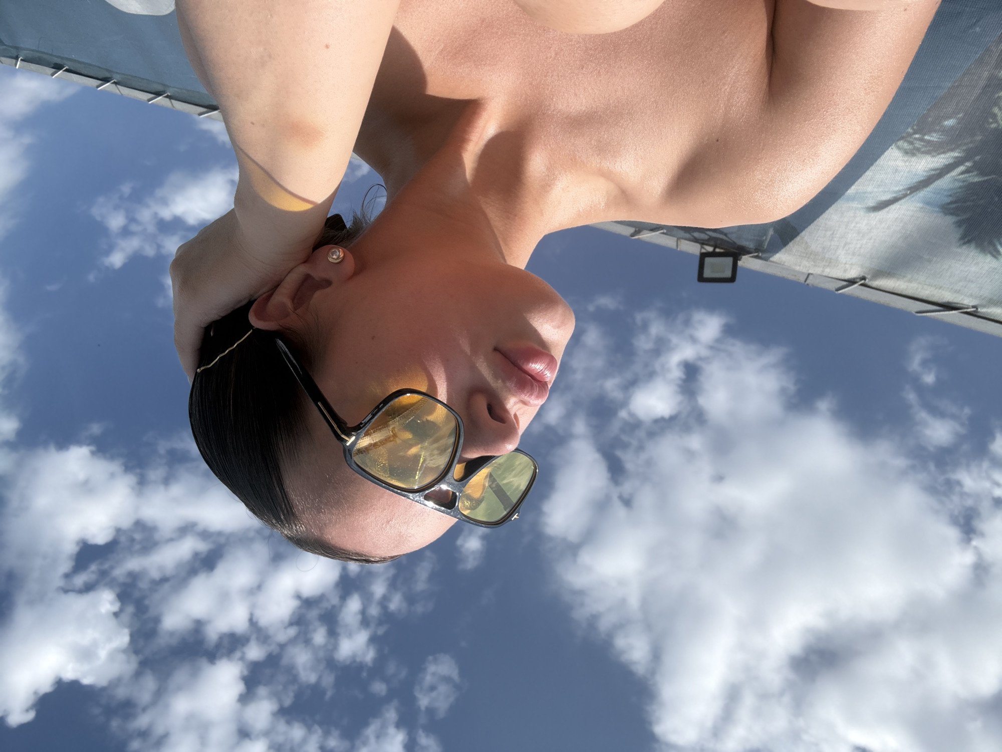 The image, taken from a low angle, shows a person with fair skin wearing sunglasses and a gold earring. The background is a blue sky with scattered white clouds. Part of the person's bare shoulder and arm are visible. Above the person's head, part of a structure with an attached light fixture and what looks like a printed fabric panel is seen. The image appears to be taken outdoors during the day. The location, Гамово, Russia, cannot be determined from the image.