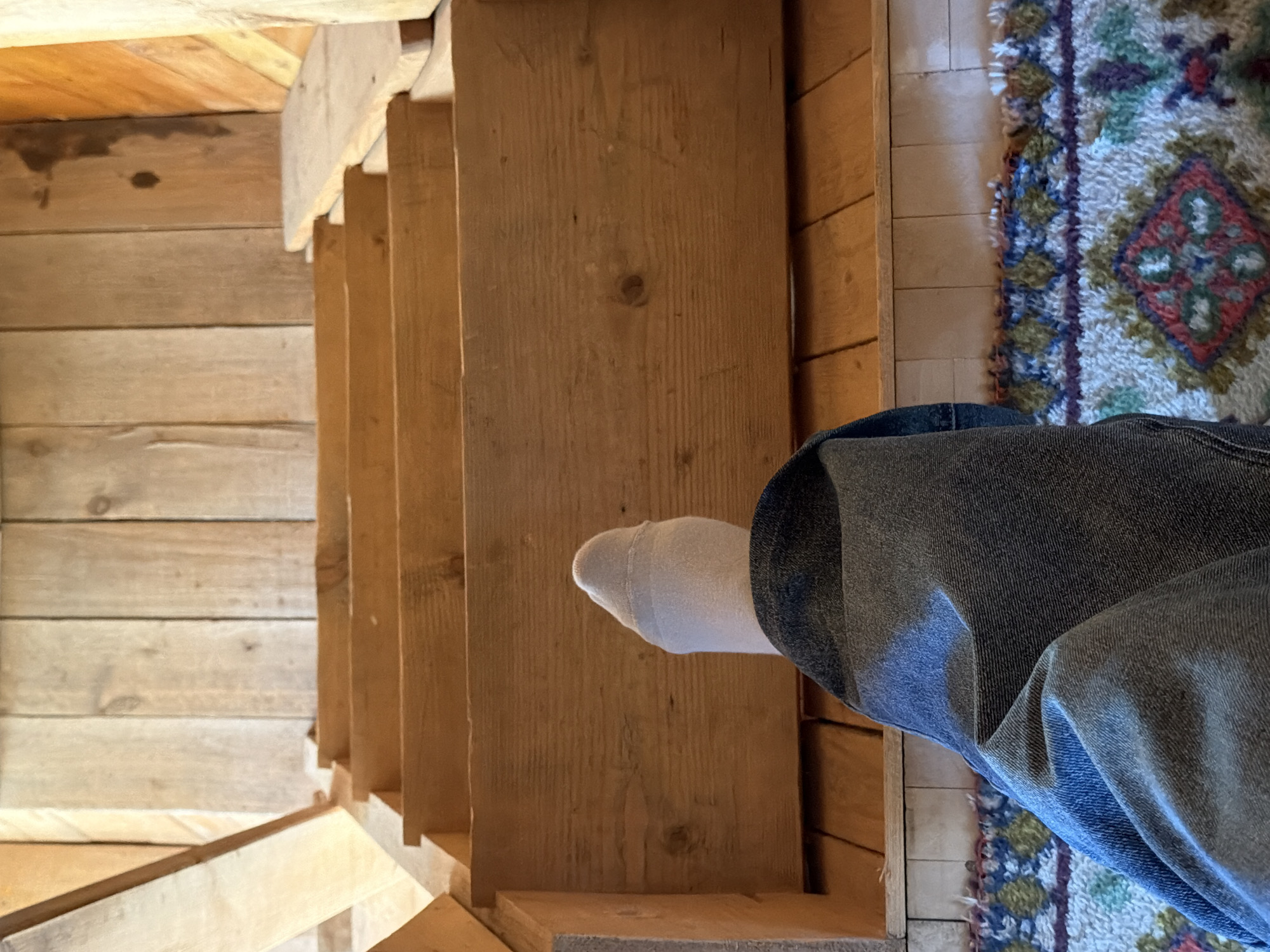 The scene depicts a first-person view of a person standing at the top of a narrow wooden staircase. The steps appear to be unfinished wood. The left wall is also constructed of unfinished wood planks. A portion of the person's leg, clad in blue jeans and a white sock, is visible on the right side of the frame. A rug with a complex pattern is present to the right of the person's leg. There is no information in the image or request that places this scene in Plamya, Russia.