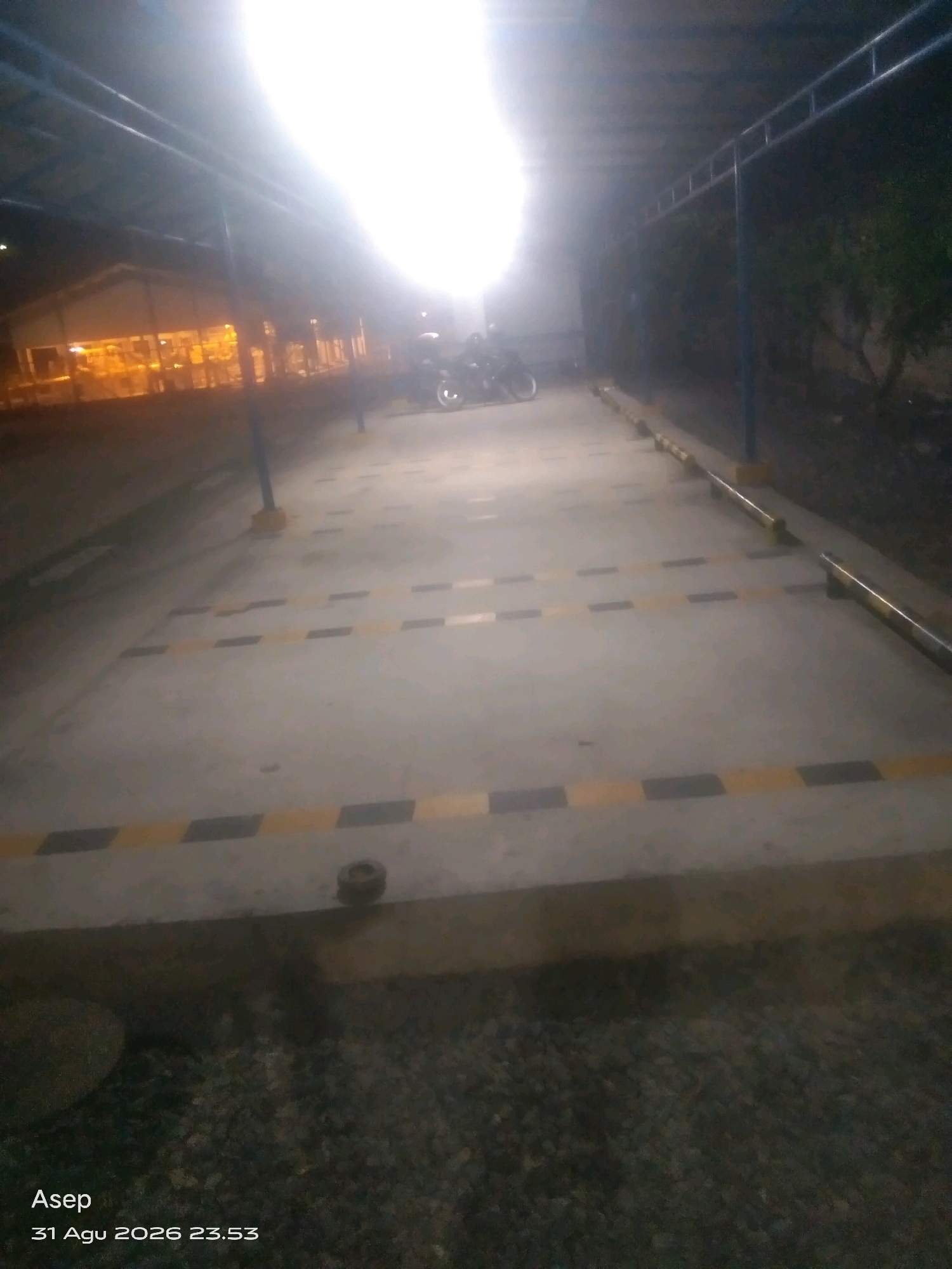 An outdoor, elevated parking lot in Way Galih, Indonesia, is captured at night. The scene is dimly lit by bright artificial lights, casting long shadows. The parking lot surface is concrete with yellow and black striped markings. A covered structure with metal beams and a roof runs along the right side of the lot, supported by blue poles. A building with illuminated windows is visible in the background to the left. A motorcycle is parked on the left side of the lot, and a person is standing near it. The foreground shows a rough, gravelly surface. The timestamp "Asep 31 Agu 2026 23.53" is visible in the bottom left corner of the image.