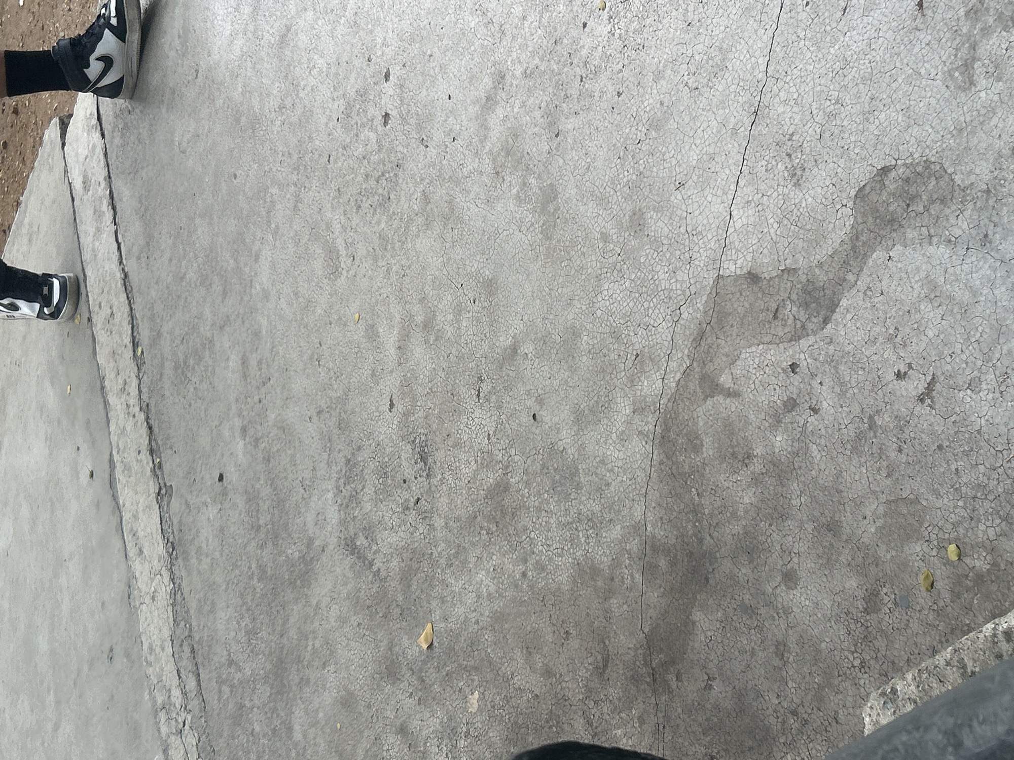 This is a ground-level, full-frame shot of a cracked, gray concrete surface. The surface shows signs of wear, with small debris and what appears to be a damp patch in the lower half of the frame. In the upper portion of the image, two pairs of feet wearing Nike sneakers are visible, suggesting the presence of at least two individuals. The environment appears to be outdoors, possibly in a public space such as a park or plaza. The lighting suggests it is daytime. The location is identified as Santa Ana, Venezuela.