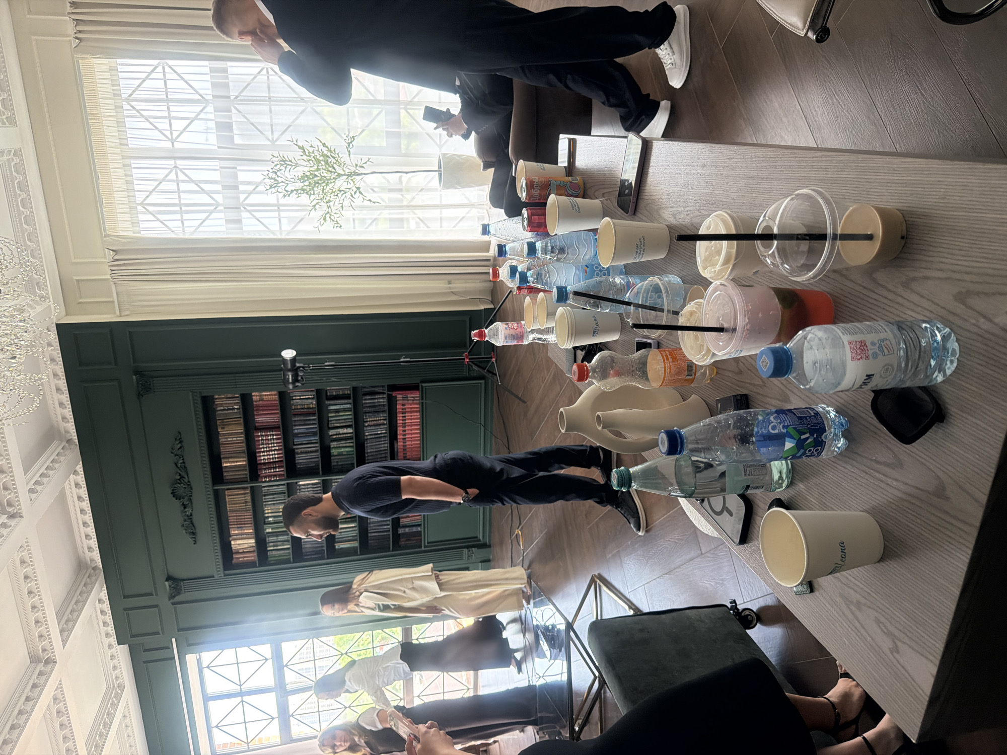 The image shows an interior setting in what appears to be a well-appointed room. A long table dominates the center, covered with numerous beverage containers, including water bottles, disposable cups, and drink canisters. A man stands near a wall with a built-in bookcase filled with books. Other individuals are visible, some holding mobile phones. One man is standing in a suit while holding his phone and is gesturing with his hand. Outside the windows, a cityscape is dimly visible. It cannot be verified from the image alone that the location is in the city of Rostov-on-Don, Russia.