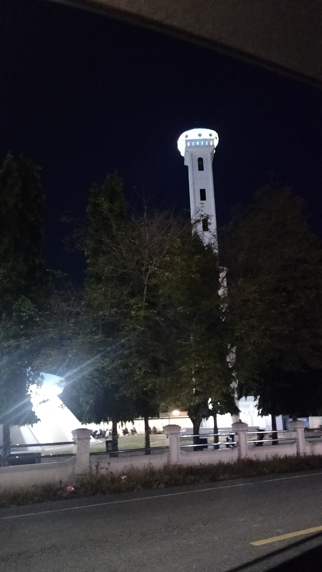 The image captures a tall, slender white tower, likely a minaret, brightly illuminated against a dark, clear night sky. The tower features small, rectangular openings and is crowned by a circular, brightly lit upper section with multiple points of light. The photograph appears to be taken from inside a vehicle, with parts of the car's interior visible at the top and bottom edges.
In the midground, several trees with varying foliage density stand between the viewer and the tower, partially obscuring it and other background structures. Below the trees, a white fence with decorative pillars runs horizontally across the frame, separating the road from a grassy area.
On the grassy area behind the fence, several motorbikes are parked, suggesting human activity or presence, possibly related to a mosque complex. Behind the trees and fence, additional brightly lit buildings are visible, consistent with a religious or public complex. A dark asphalt road with a faint yellow line is visible in the foreground. The time of day is clearly night, as indicated by the dark sky and the artificial illumination of the structures. The location is Lambarotunong, Indonesia. No discernible text is visible in the image.