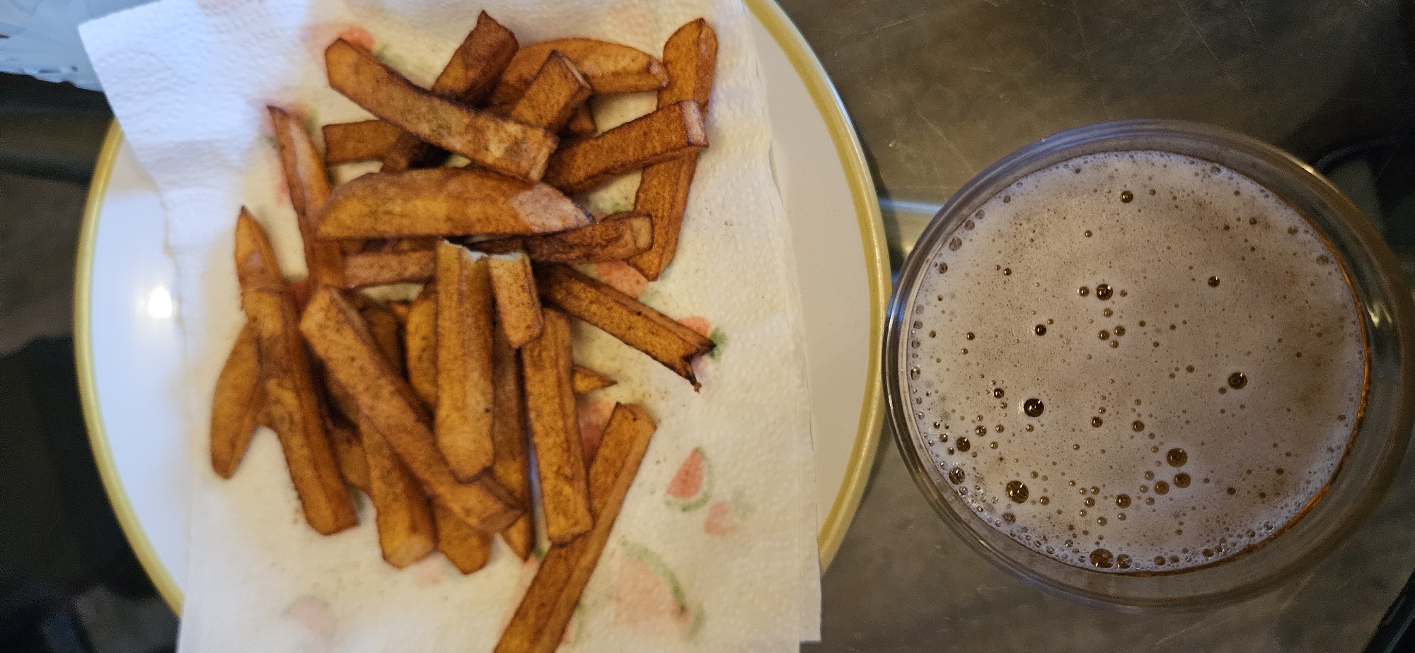 The image presents an overhead shot of a casual meal or snack, featuring a plate of chips and a drink, set on a dark, reflective surface.
On the left, a white plate with a prominent yellow rim holds a generous portion of golden-brown, thick-cut chips (fries). These chips are laid out on a white paper towel, which has faint, pastel-colored designs resembling watermelon slices in pink and green. The chips appear freshly cooked, with a crispy texture and varying shades of brown, indicating they are well-fried. They are likely placed on the paper towel to absorb excess oil.
To the right of the plate, partially cropped, is a clear glass containing a dark beverage. The drink is topped with a substantial layer of light-brown foam, dense with numerous small bubbles, typical of a beer such as an ale or stout, or potentially a dark carbonated soft drink.
The background surface appears to be a dark, smooth countertop or table, possibly metallic or stone, reflecting some ambient light. The lighting suggests an indoor setting, possibly evening. No people are visible, and there is no discernible text within the image. The scene depicts a simple, comforting food and drink pairing, common in a relaxed, home-like environment in Norwich, United Kingdom.
