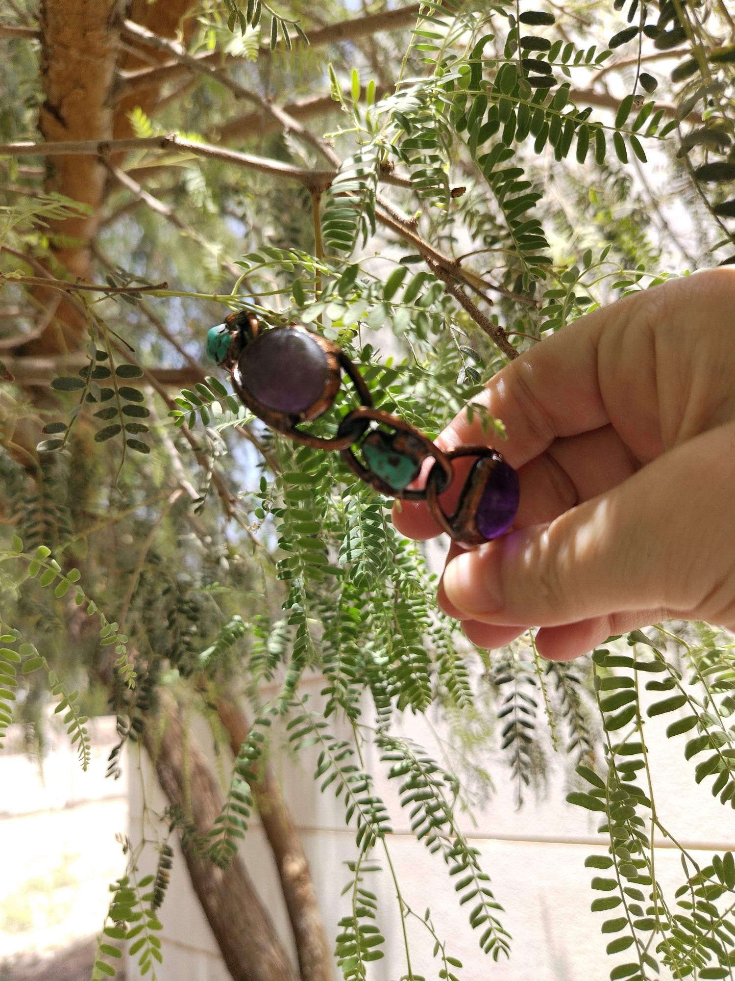 A hand holds a pair of gemstone-embedded eyeglasses in front of a mesquite tree. The glasses have dark purple and turquoise gemstones. The background is dominated by the green foliage of the tree. The photo was taken in the city of Al Mas'oudi, United Arab Emirates.