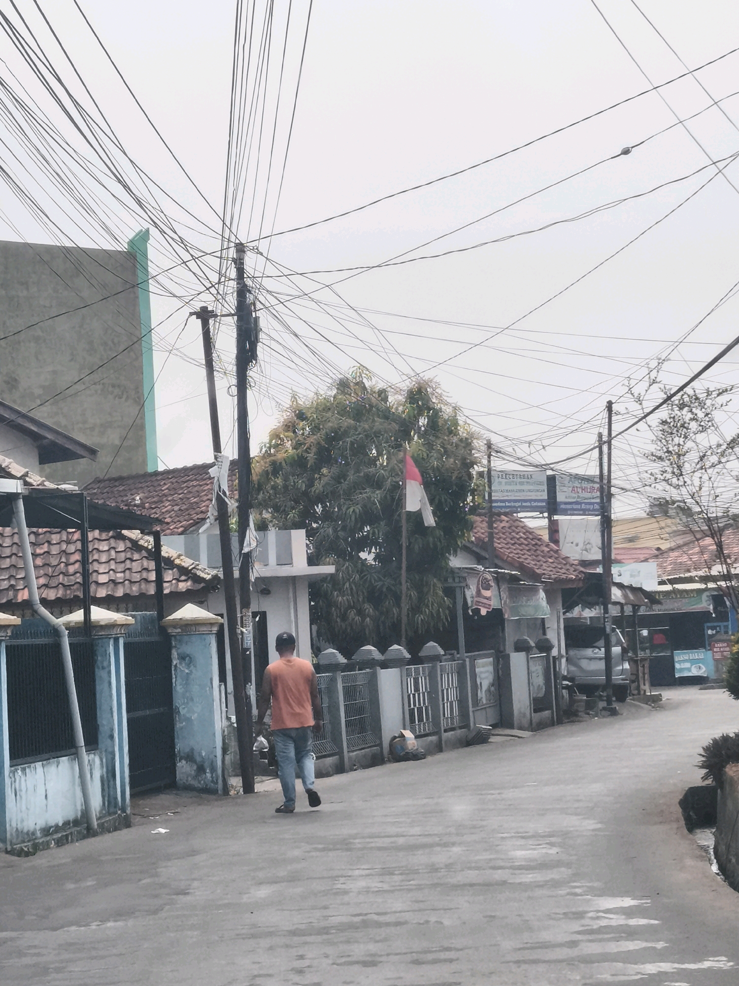 The image depicts a street scene in Palembang, Indonesia, characterized by a dense network of overhead electrical wires. In the foreground, a person wearing an orange t-shirt and blue jeans walks away from the camera on an asphalt road. To the left of the road, a blue and grey fence with decorative pillars borders a property with a tiled roof. Across the street, a cluster of buildings with red-tiled roofs is visible, along with trees and more electrical poles. Several signs are mounted on poles in the middle ground, including one with the Indonesian flag. The overall impression is of a residential or commercial street in a densely populated area, possibly experiencing overcast weather due to the washed-out sky.