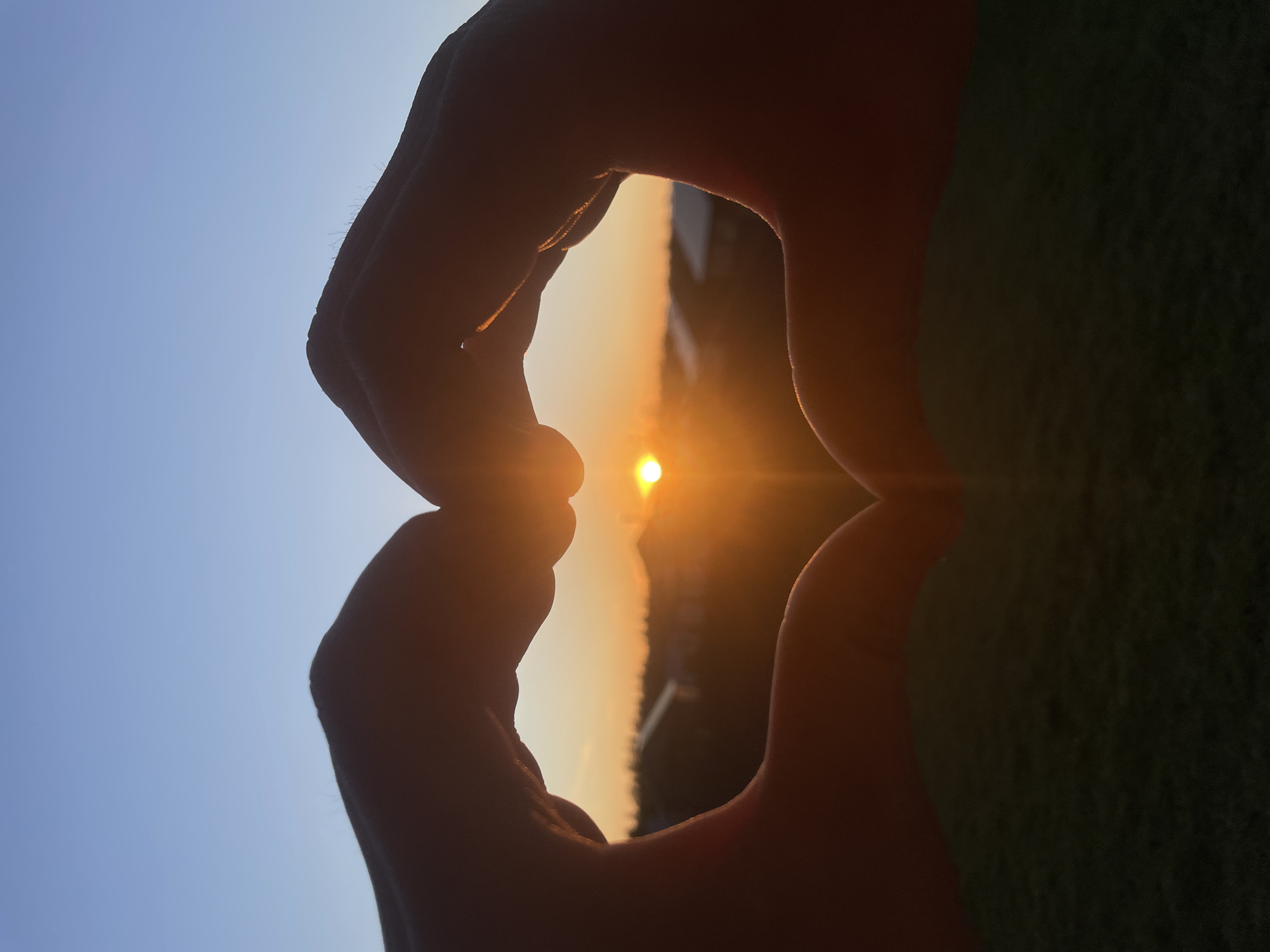 The image shows a close-up of two hands forming a heart shape with a sunset visible through the opening. The background includes a view of trees, buildings, and sky. The location is Підшивалово, Russia. The time of day is sunset.