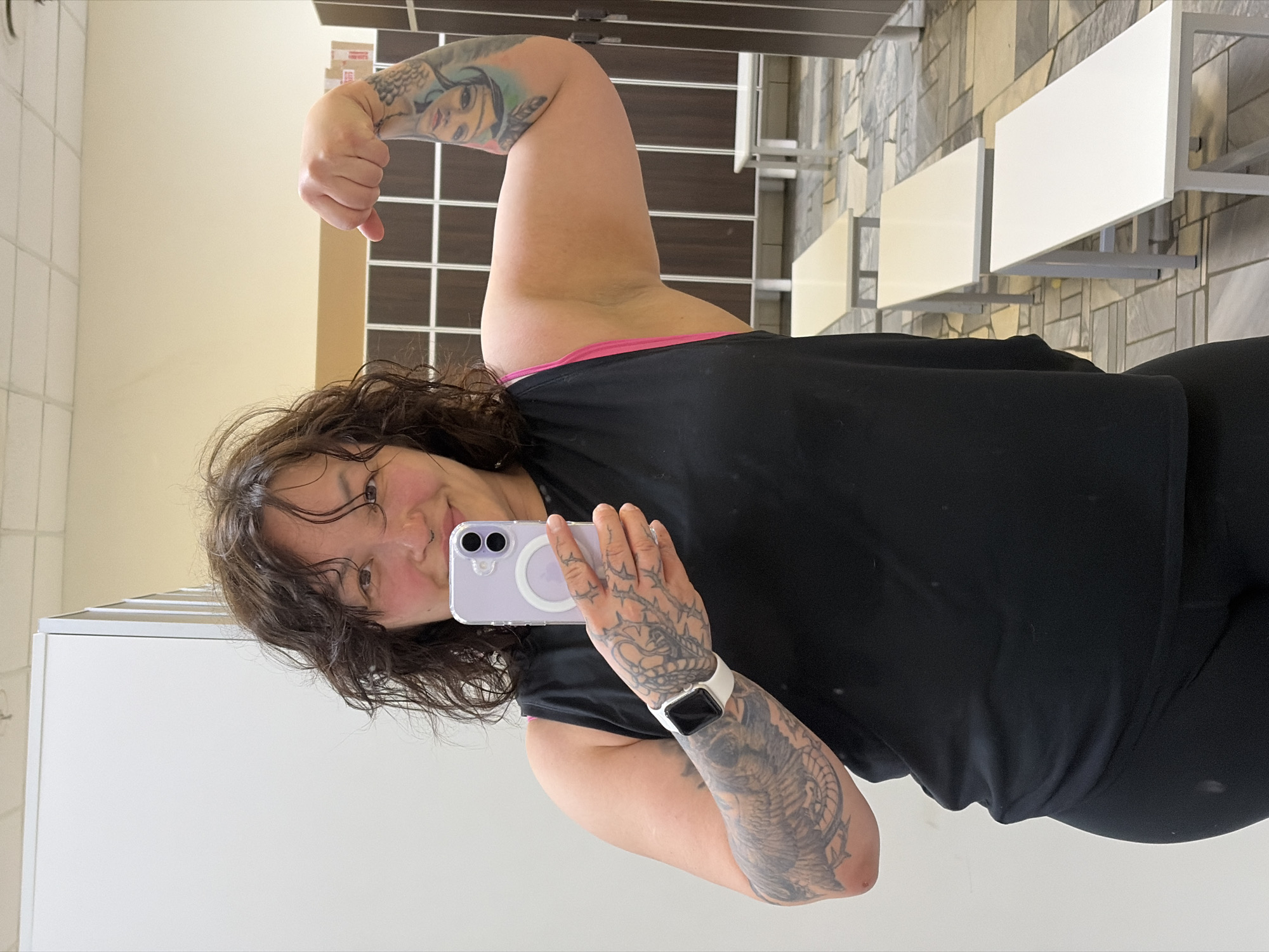 In a locker room, a woman with dark, curly hair and a nose ring takes a selfie in a mirror. She is wearing a black tank top, black leggings, and a white Apple Watch. Her left arm is flexed, and she has tattoos on both arms. In the background, there are rows of white benches and wooden lockers. The floor is tiled. The image does not provide any specific visual cues to determine the exact location within the city of Chaikovsky, Russia.